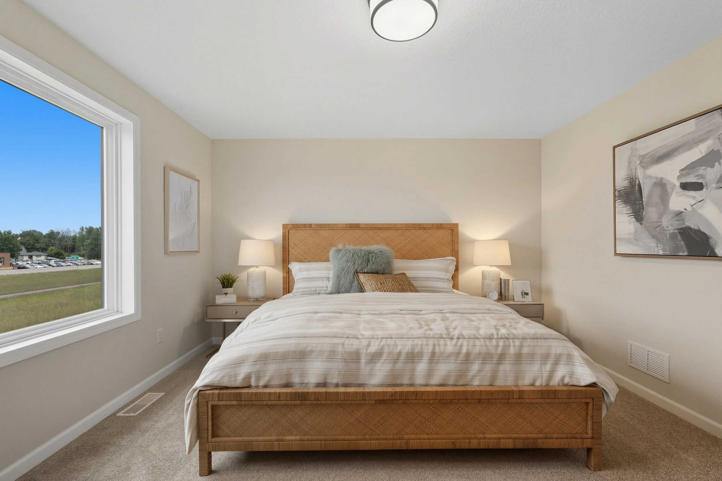 A bedroom with a large bed, two nightstands with lamps, and a large window showing a grassy landscape outside.