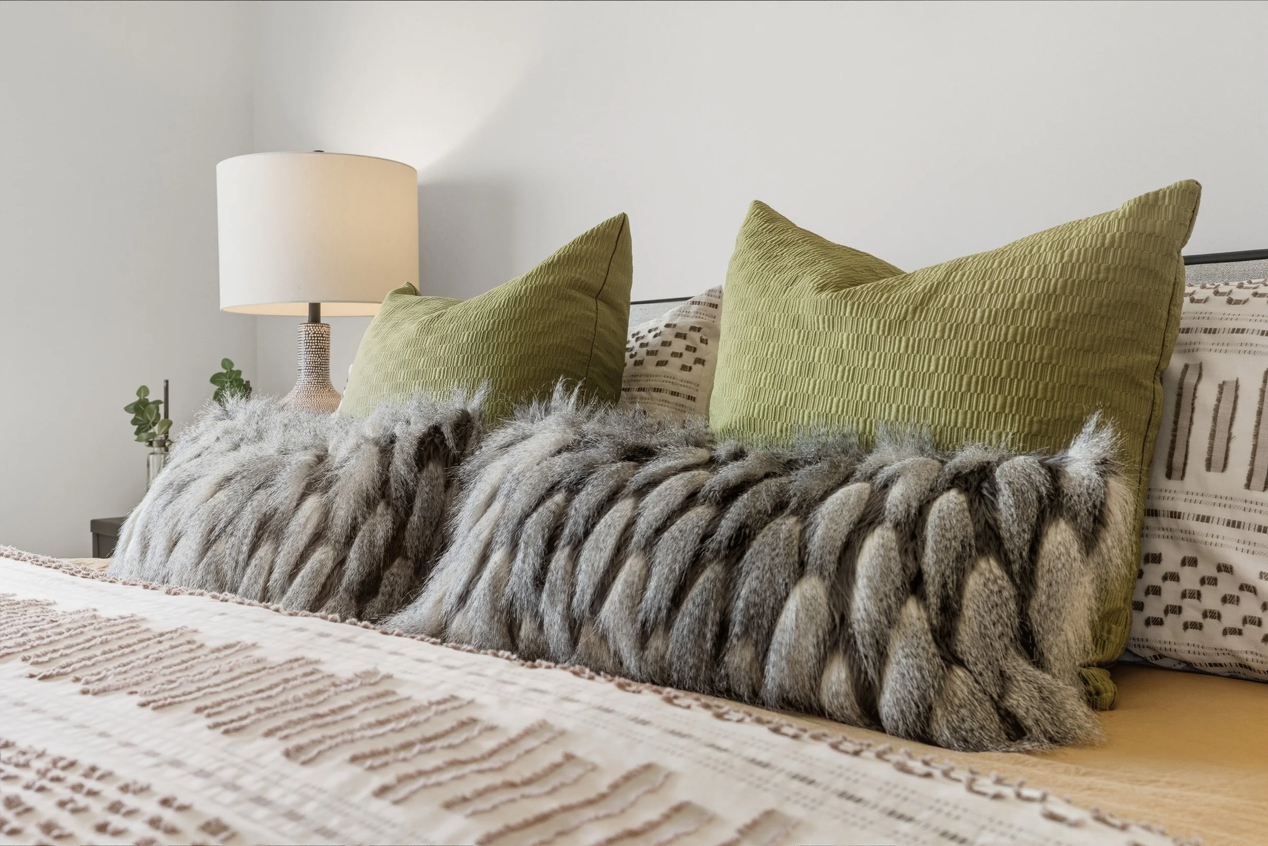 Decorative bed with two green pillows, a faux fur throw, and patterned bedding in a modern bedroom with a table lamp and small plants on bedside tables.