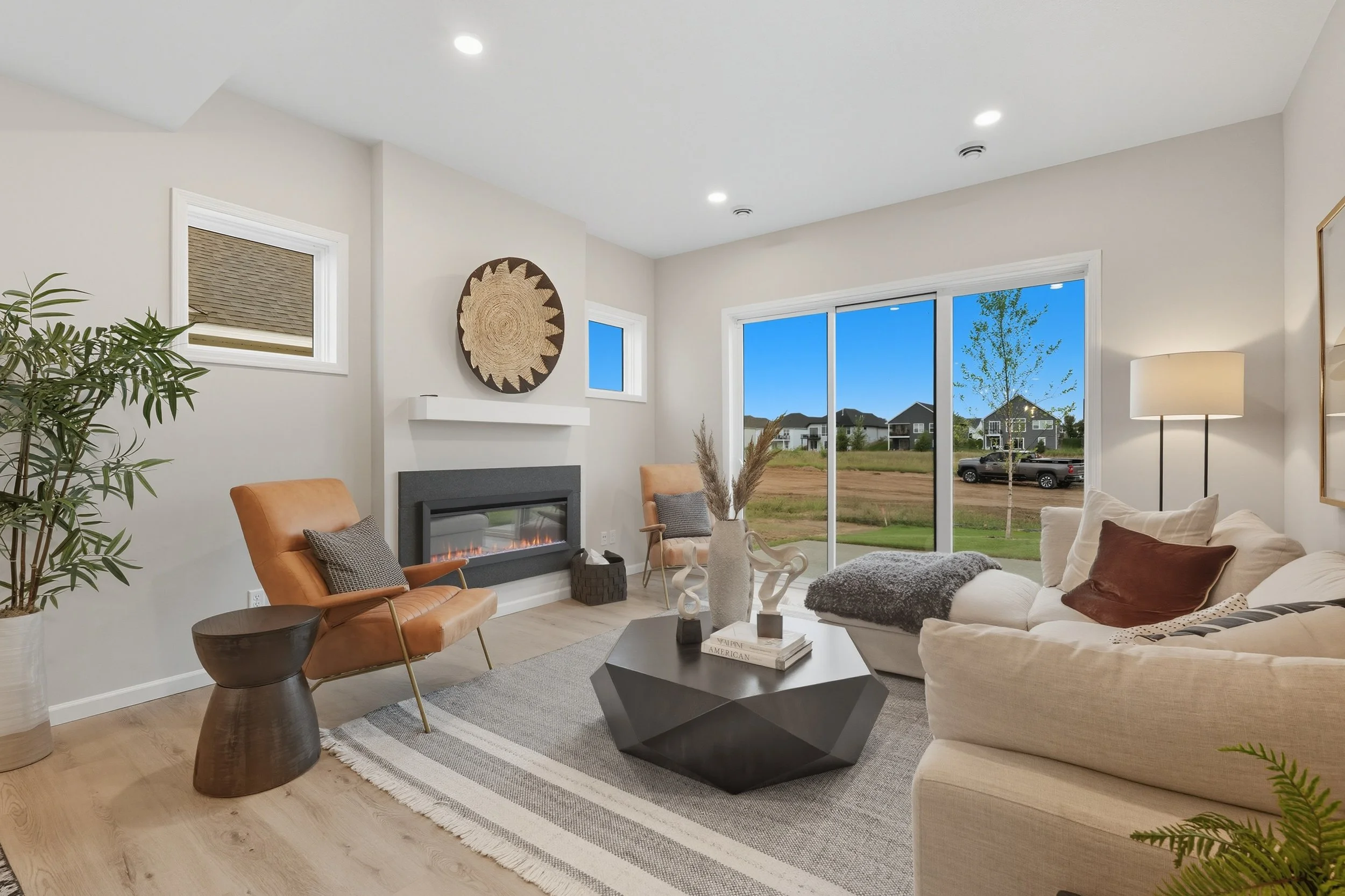 Modern living room with beige sofas, a black geometric coffee table, a tan leather chair, a fireplace, large sliding glass doors showing an outdoor scene, and home decor including art and plants.