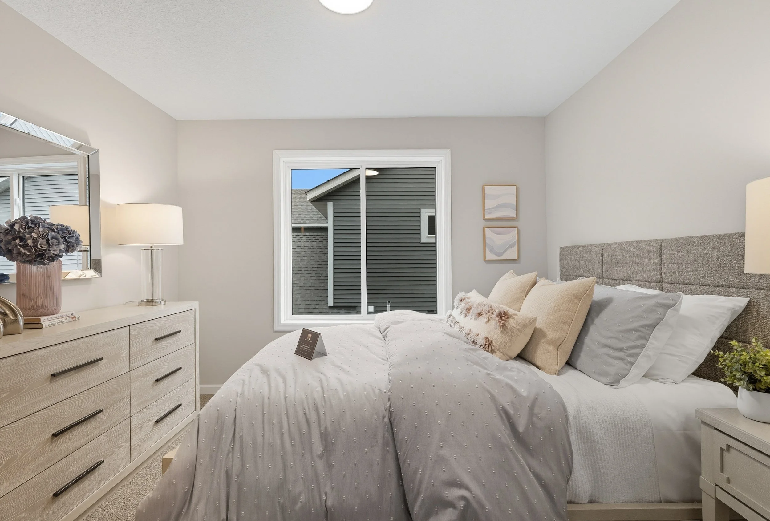 A bedroom with a bed, dresser, and window showing a neighboring house's exterior siding