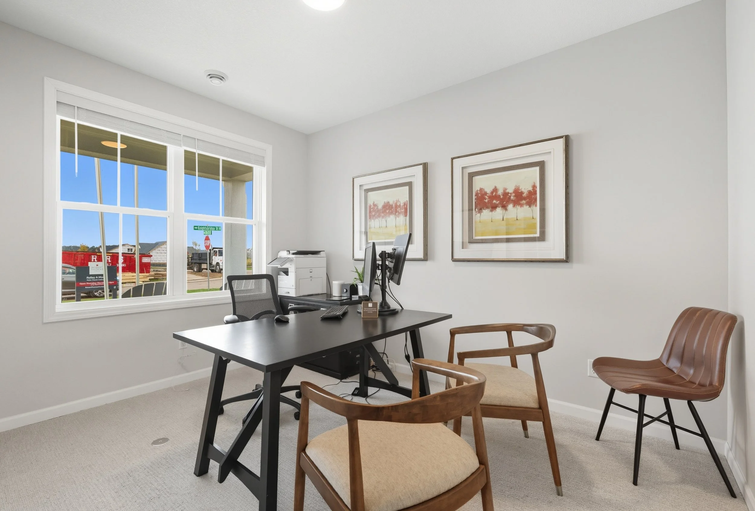 Modern office room with white walls, black desk, ergonomic chair, two wooden chairs, framed artwork of trees, and large window with view of a suburban street.