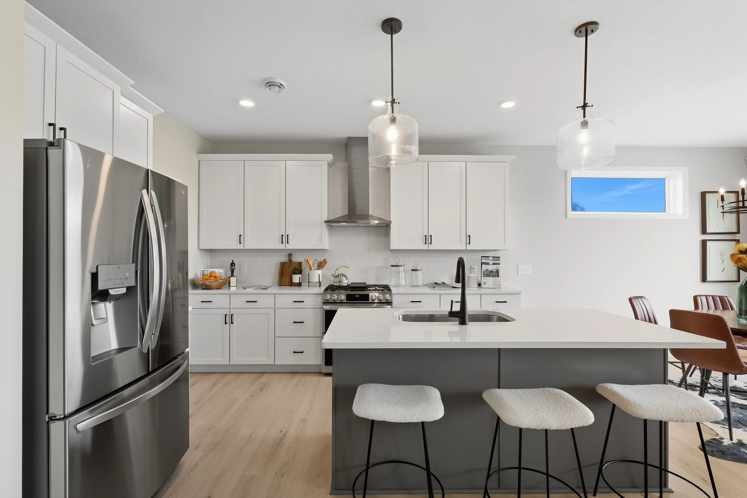 Modern kitchen with white cabinets, stainless steel refrigerator, island with black faucet and beige stools, small window with blue sky, and a dining area with brown chairs.