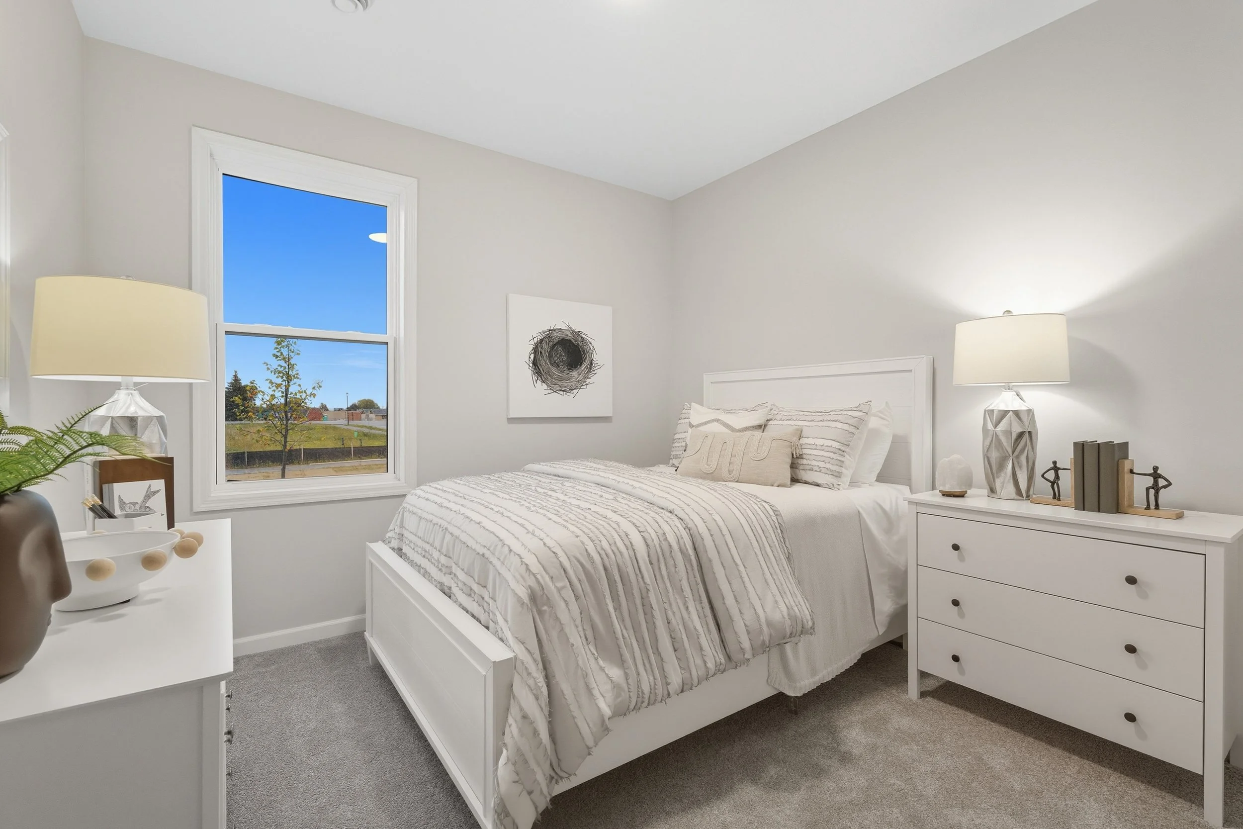 A bedroom with white walls, a white bed with striped bedding, a window showing a blue sky, and white furniture including a dresser with a lamp and decorative items.