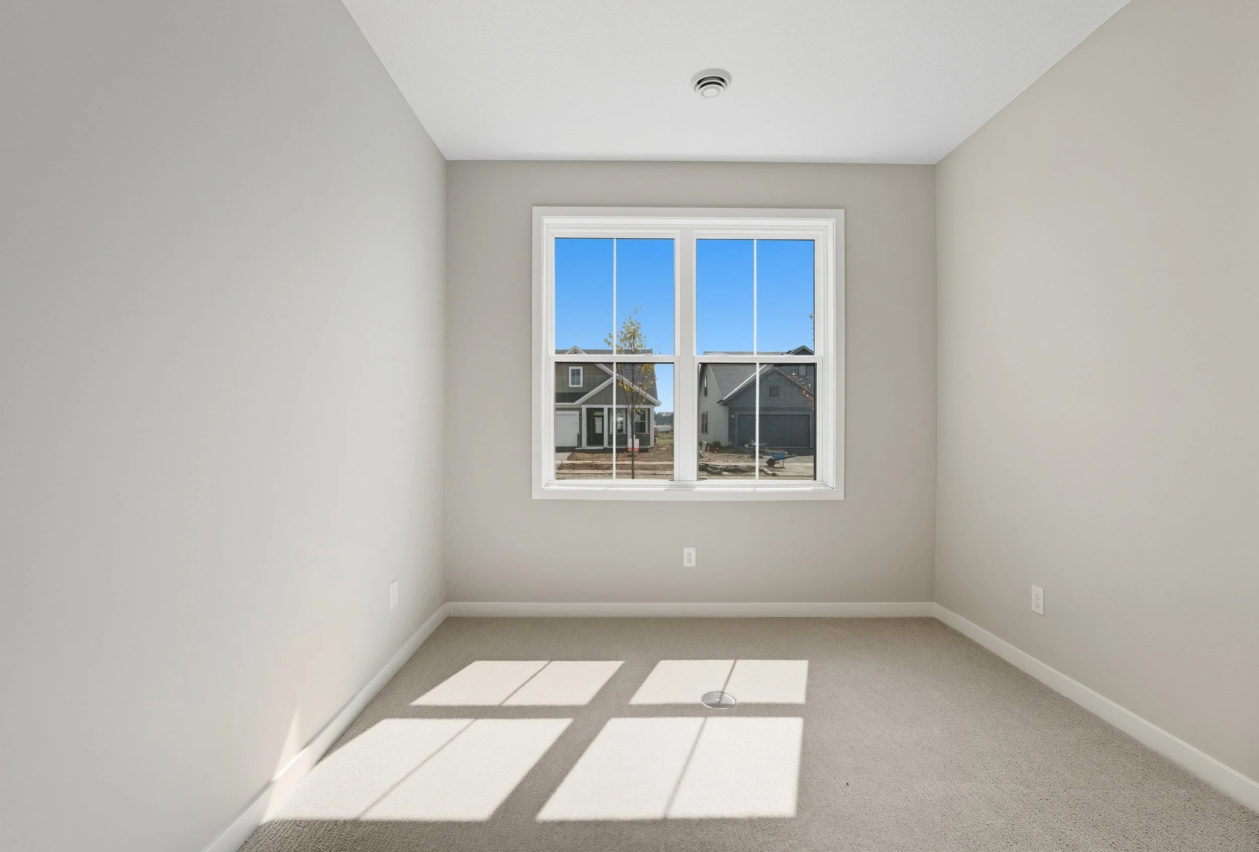 Empty room with beige carpet, white walls, white ceiling, and a window showing a view of neighboring houses and a clear blue sky.