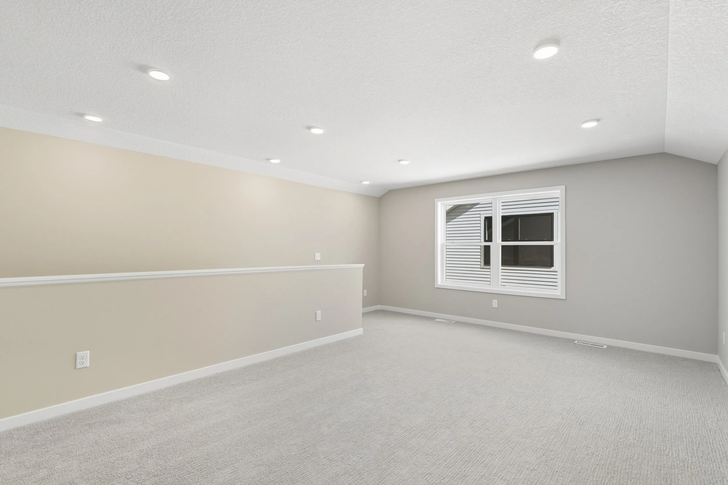 Empty beige and gray room with carpeted floor, white baseboards, and ceiling lights, featuring a window with blinds.