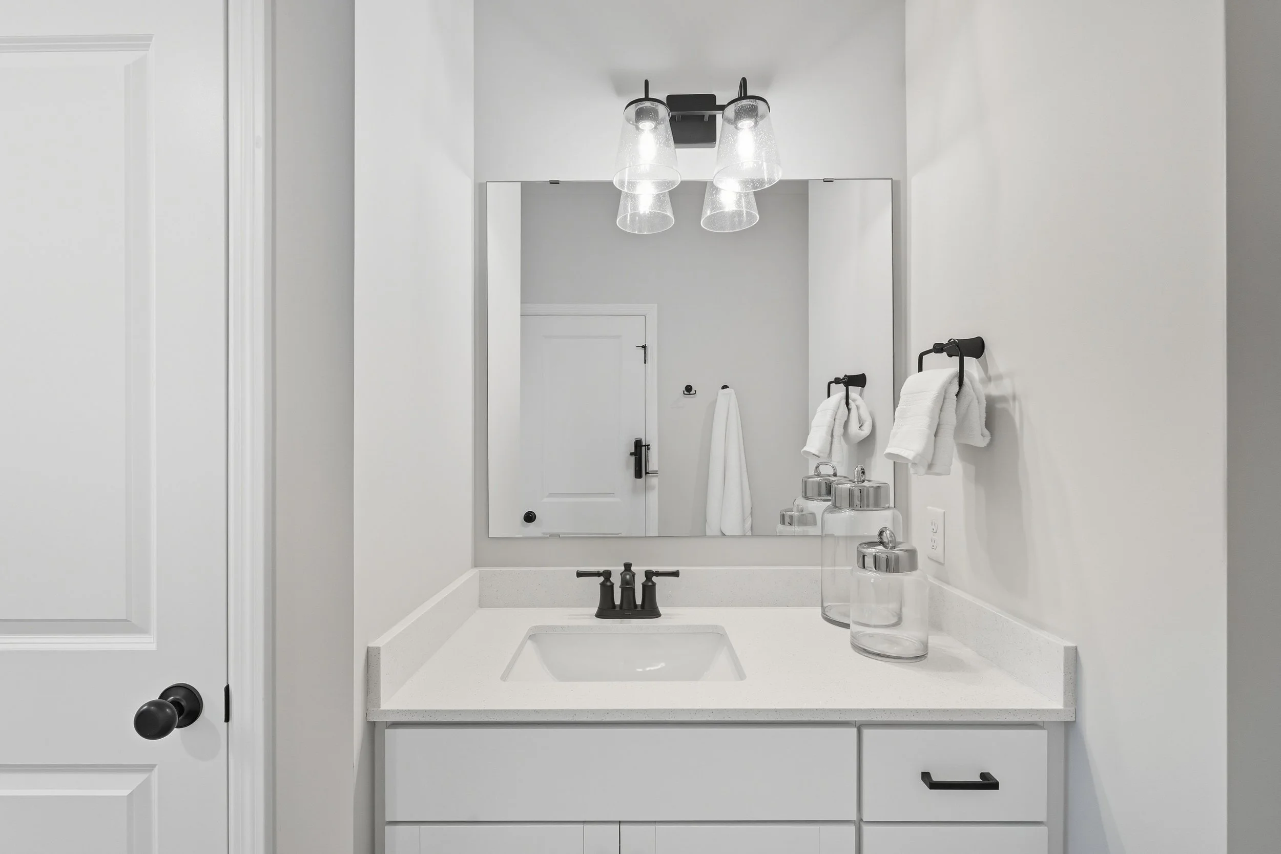 Minimalist bathroom with white vanity, black faucet, large mirror, and modern light fixture, with white towels and glass containers on the sink counter.