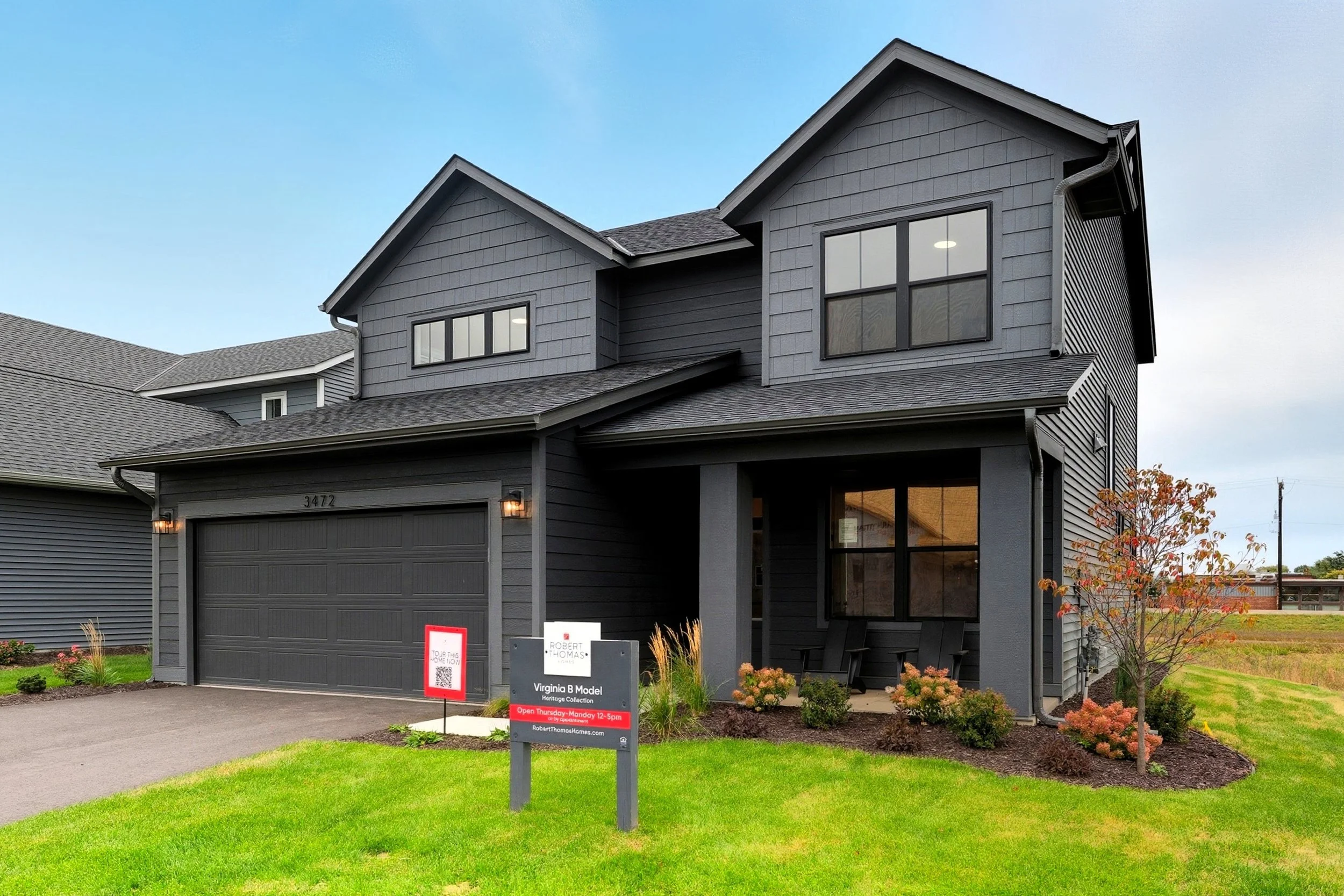 A two-story gray house with black trim, a two-car garage, and a well-maintained lawn with small shrubs and a young tree in front.