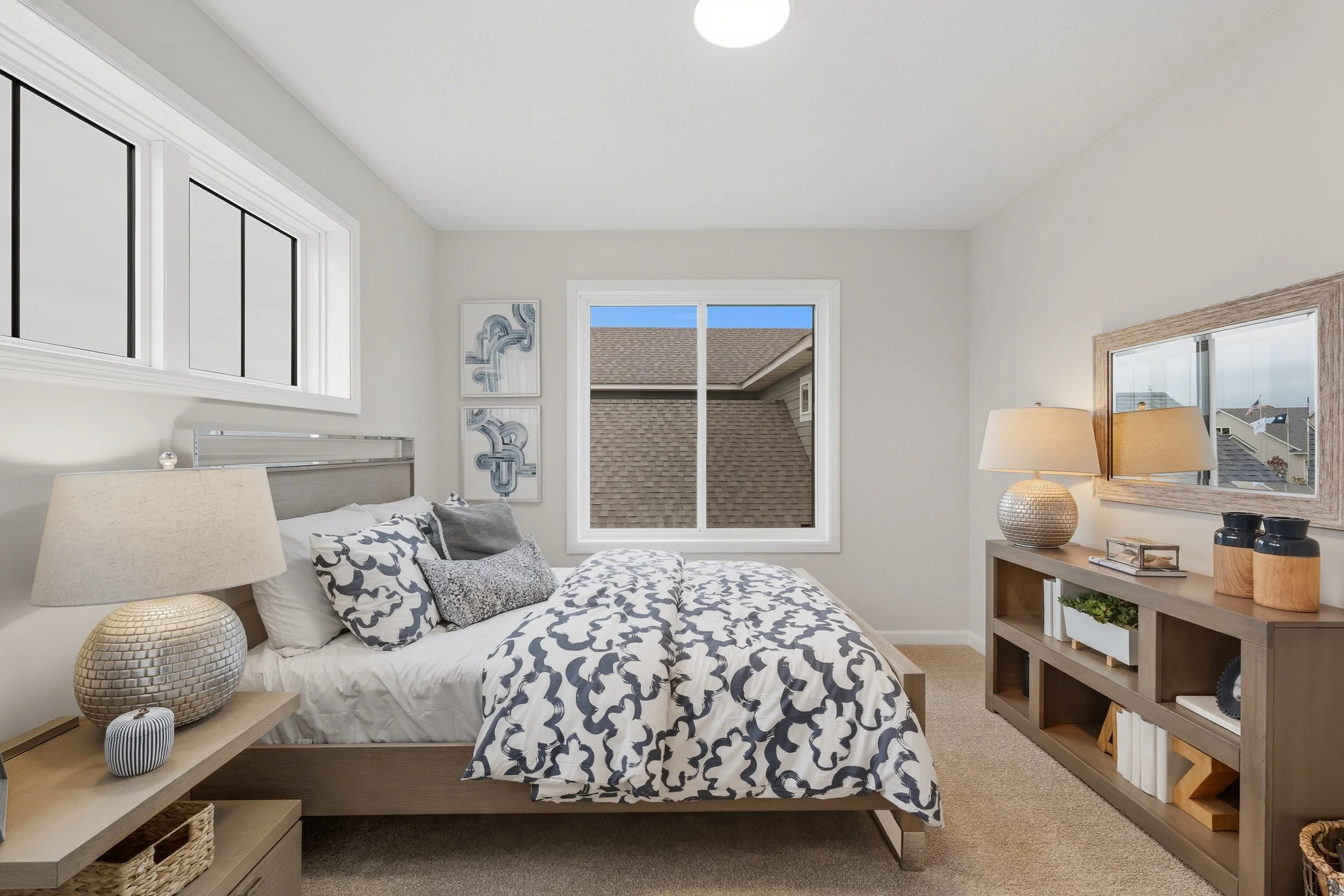 Bedroom with a bed, decorative pillows, a nightstand with a lamp, a large mirror, and a wooden shelving unit with lamps, books, and decorative objects.