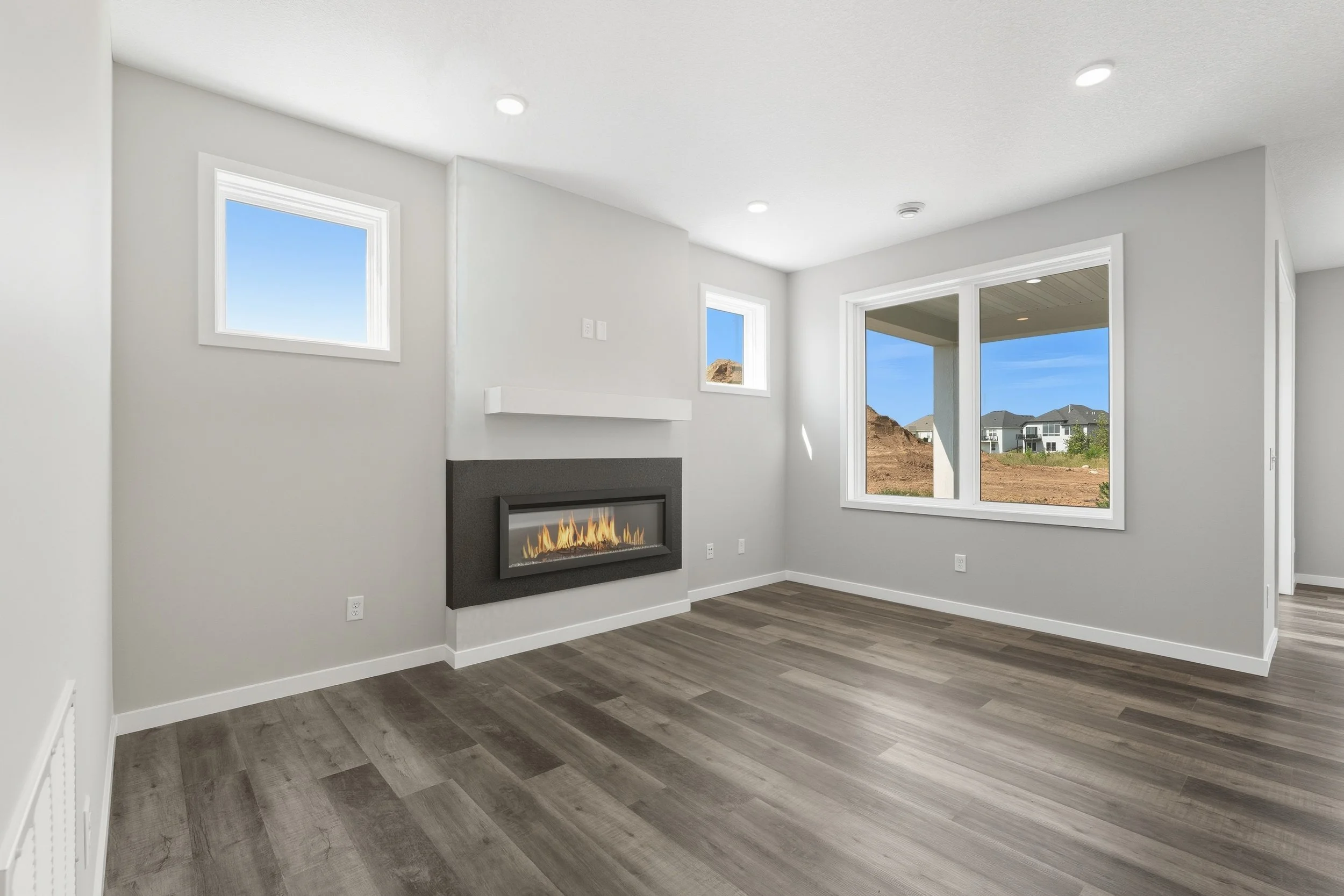 Empty living room with gray flooring, white walls, a wall-mounted fireplace, two small windows, and large sliding glass door leading to a backyard with houses in the distance.