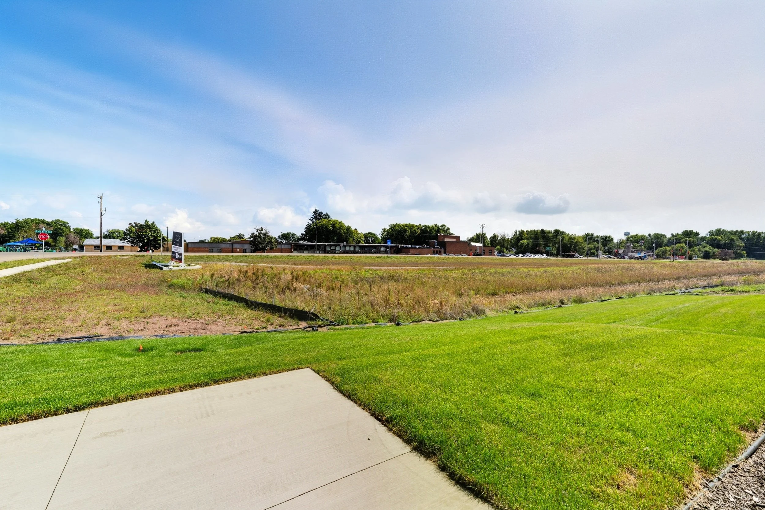 Wide view of a grassy area with a sidewalk, developing field, and a building in the distance under a partly cloudy sky.