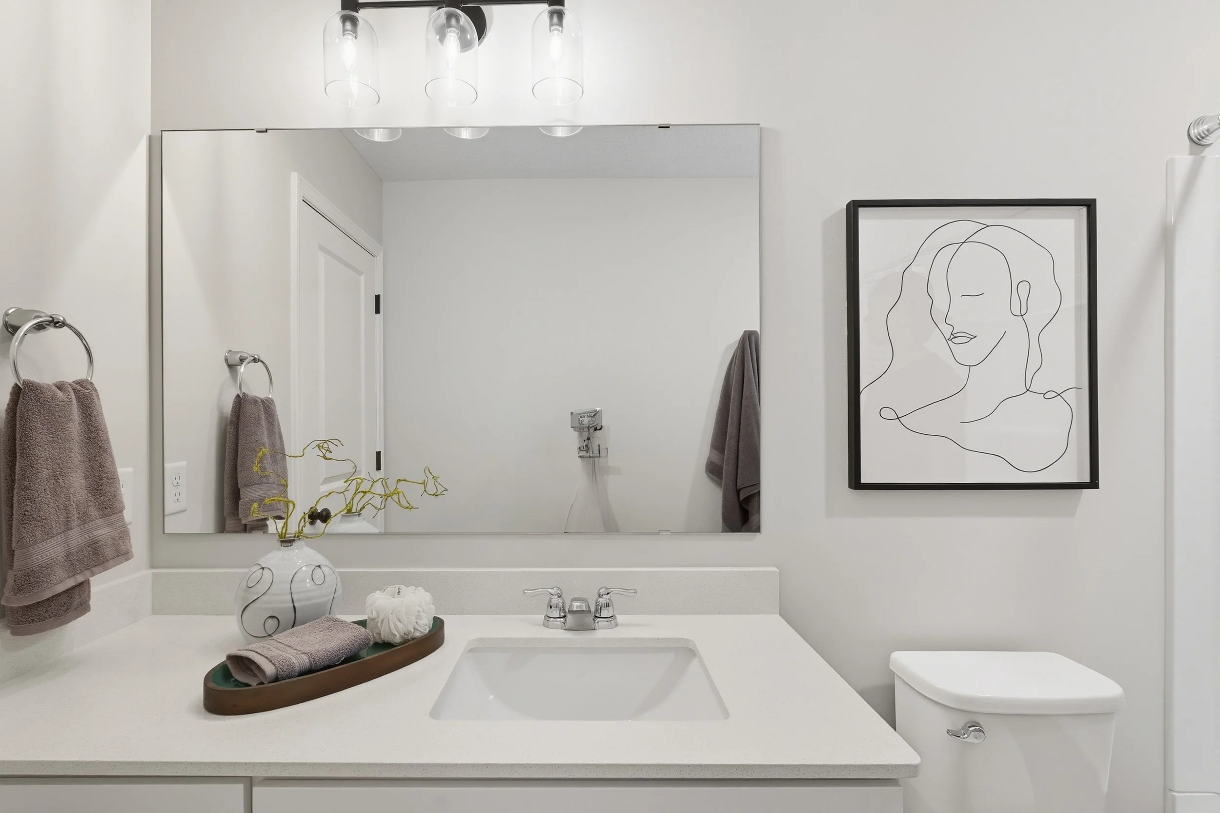 A modern bathroom with a large mirror, white countertop with a sink, gray towels on towel rings, a decorative vase with twisted branches, framed line art of a woman's face, and toilet.
