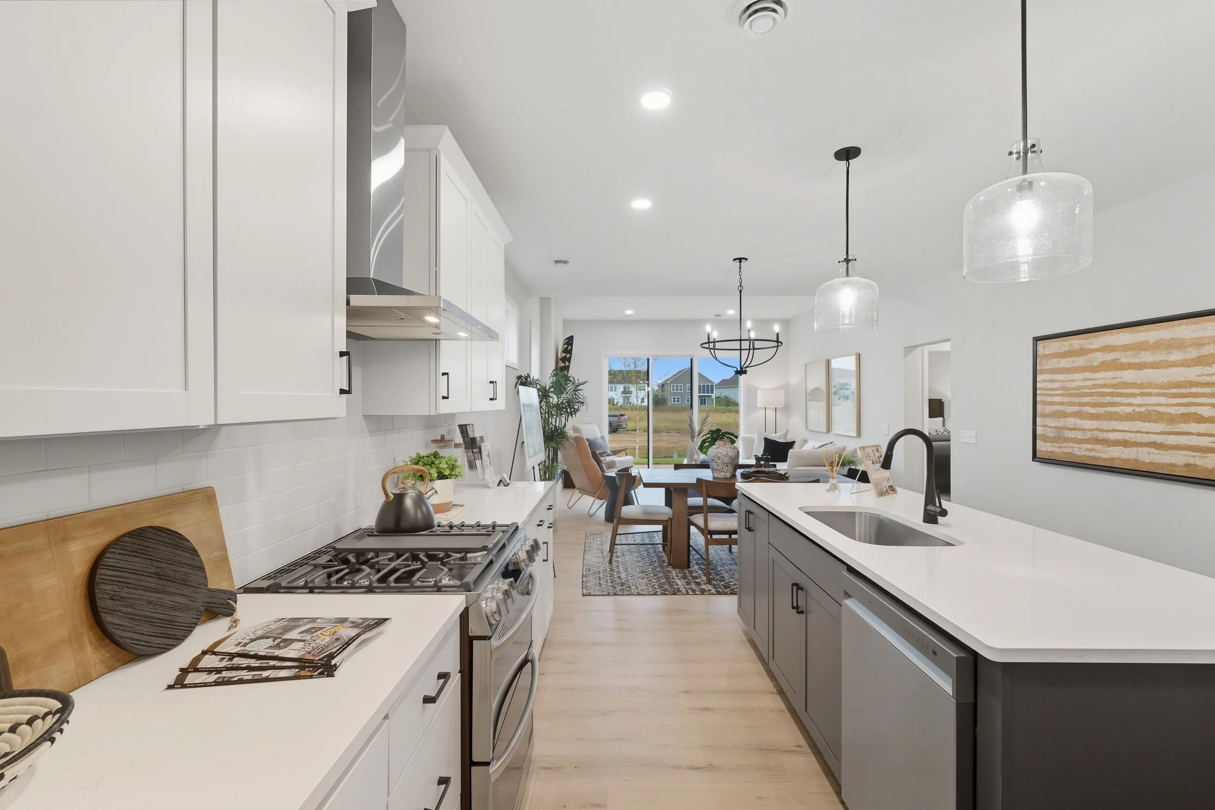 Open-concept modern kitchen with white upper cabinets, gray lower cabinets, stainless steel appliances, a large kitchen island with a sink, and a dining area with a table and chairs, leading to a living room with a view of outdoor houses through larg