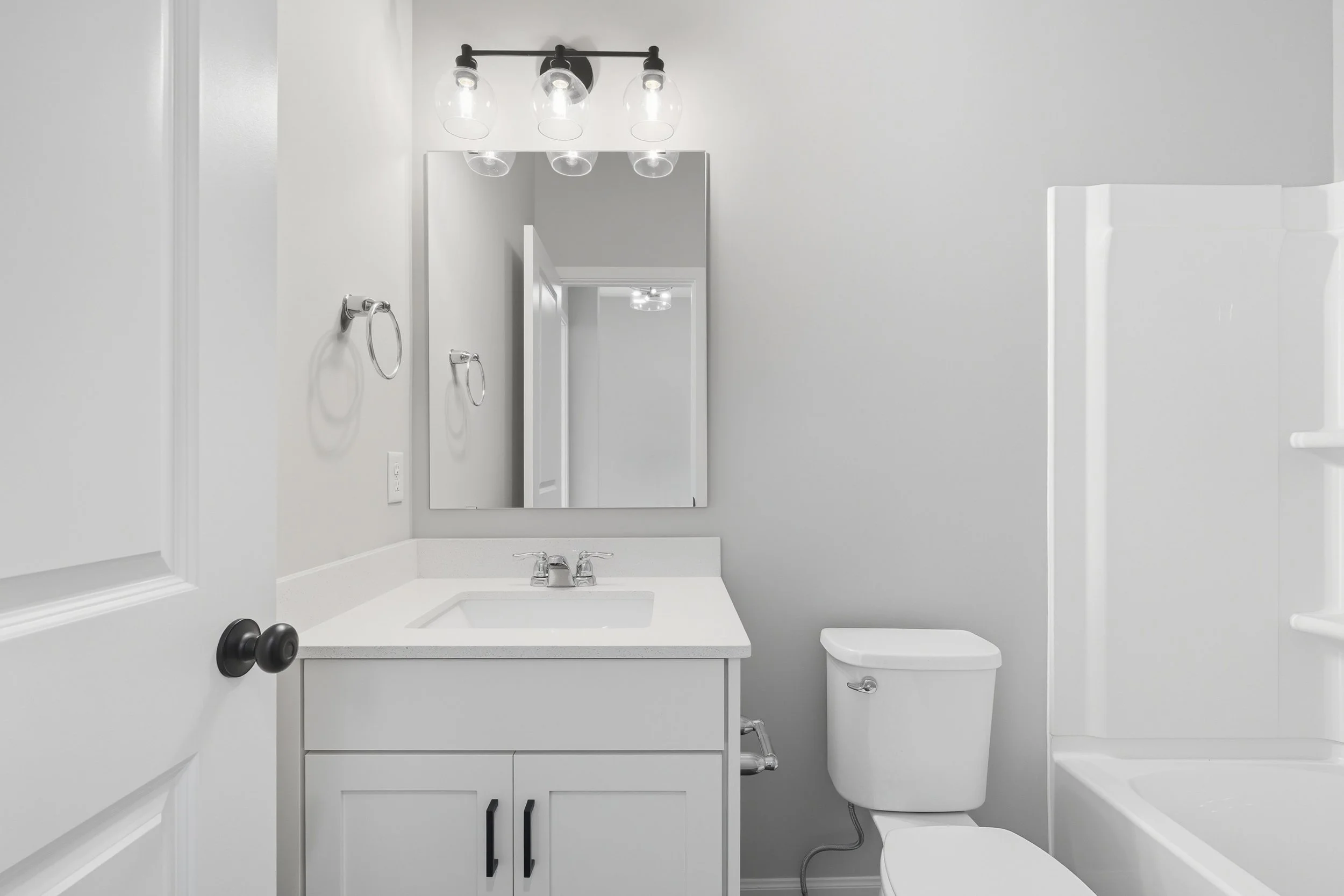 A minimalist bathroom with a white vanity, a mirror, a toilet, and a shower stall, illuminated by three glass light fixtures above the mirror.