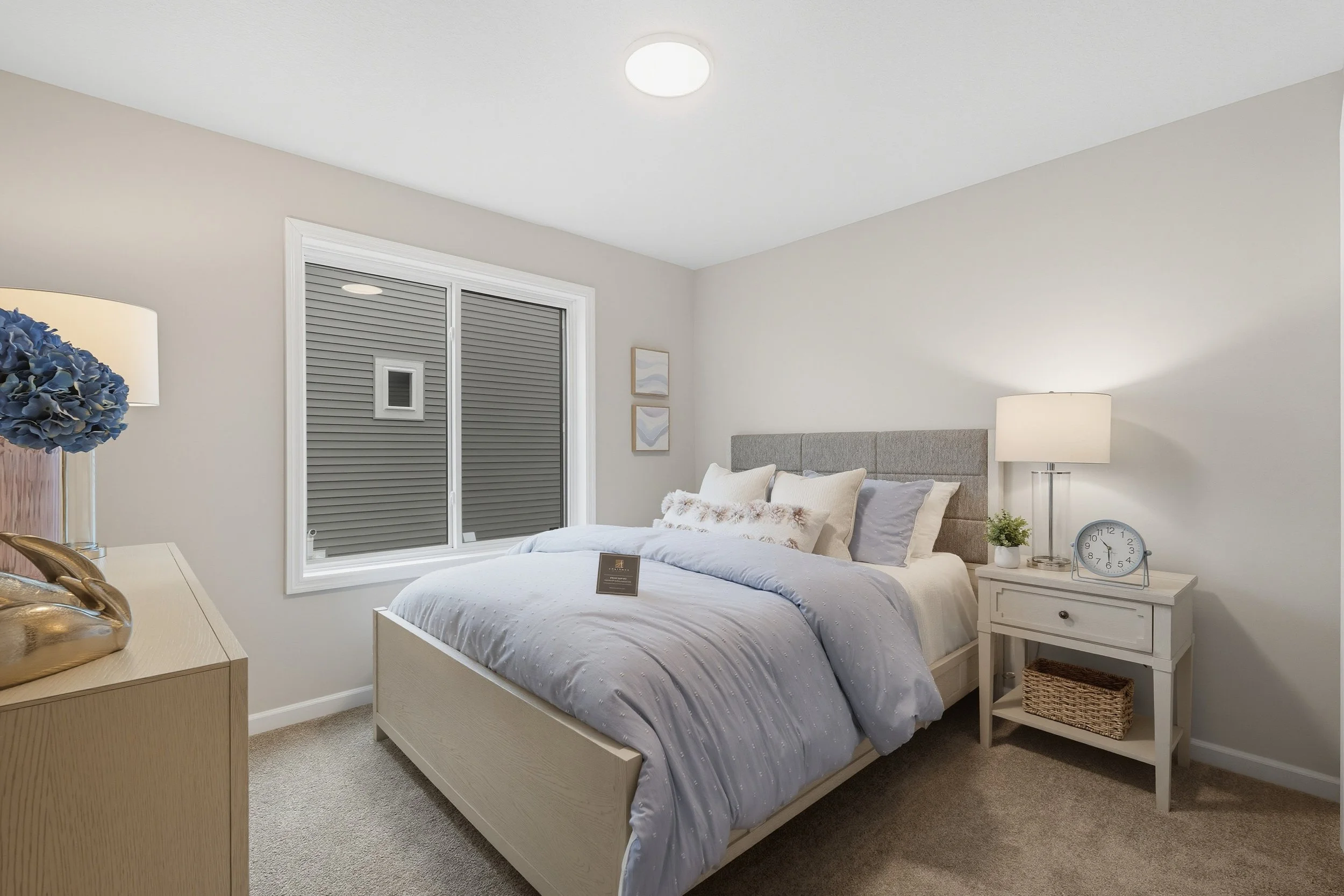 A neatly made bedroom with a bed, nightstand, lamp, clock, and decorative pillows, with a window covered by gray blinds and minimal wall art.