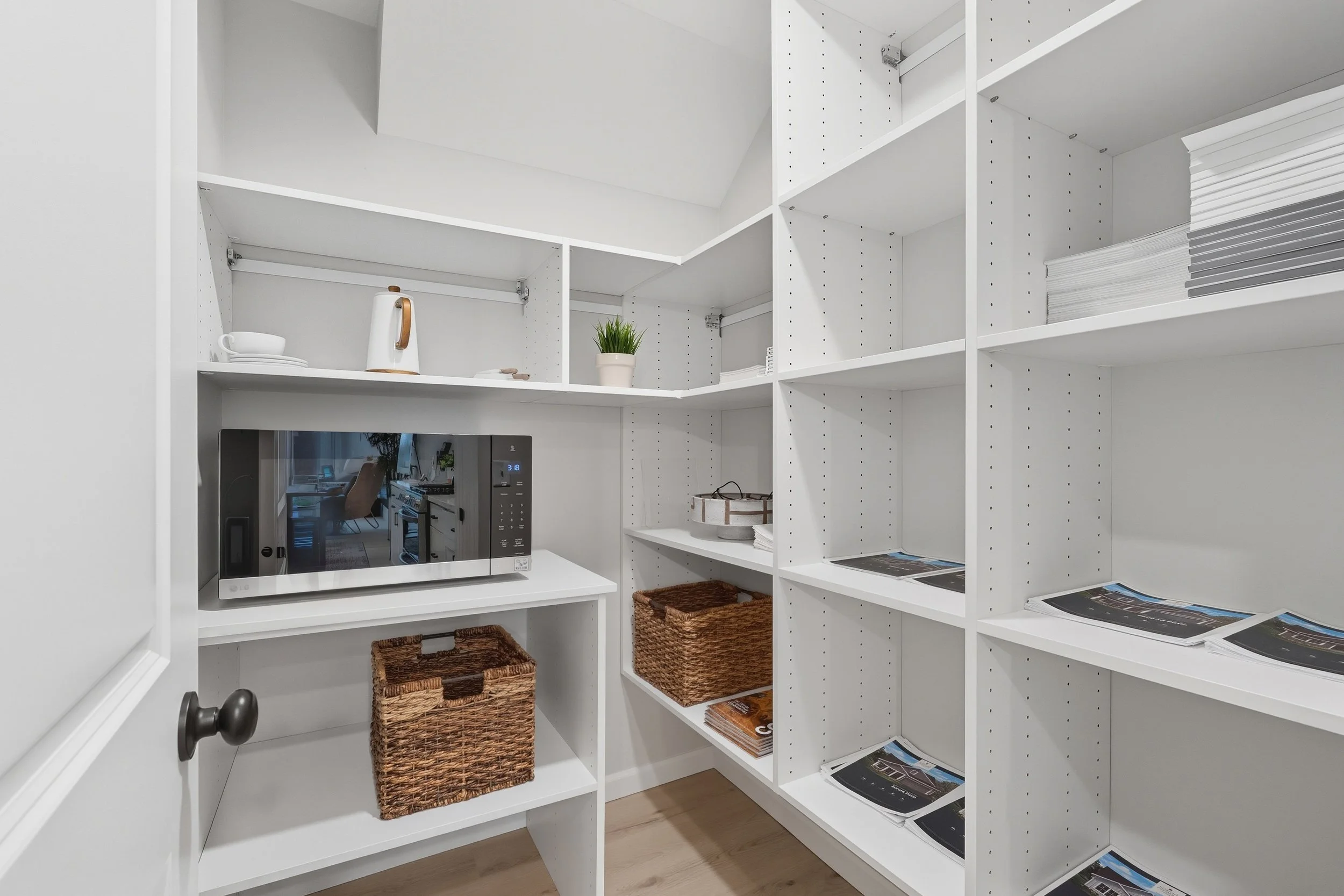White organized pantry with shelving, wicker baskets, a microwave, and some paperwork.
