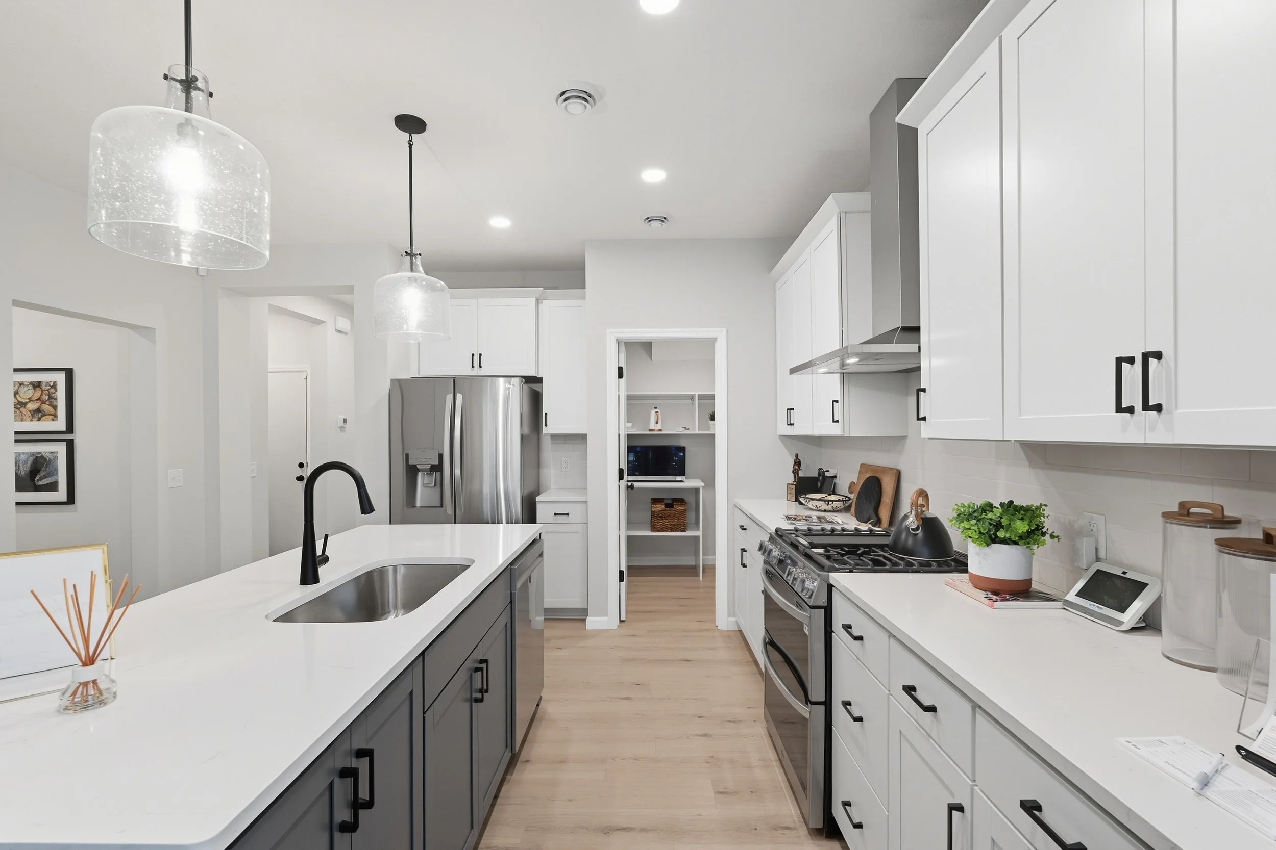 Modern kitchen with white cabinets, stainless steel refrigerator, black handles, black faucet, white countertops, and light wood flooring. Decor includes a potted plant, cutting boards, and small appliances.
