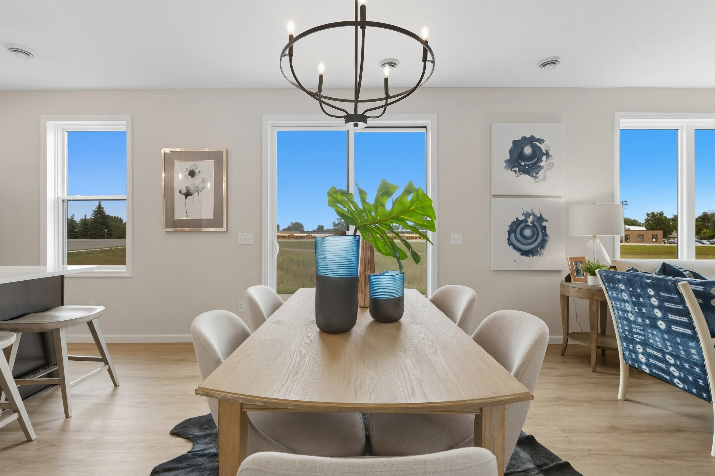 A modern dining room with a light wood table, surrounded by cream-colored chairs, with three blue vases and a green plant centerpiece. The room has three windows with a view of green grass and trees, an abstract artwork on the wall, and a chandelier