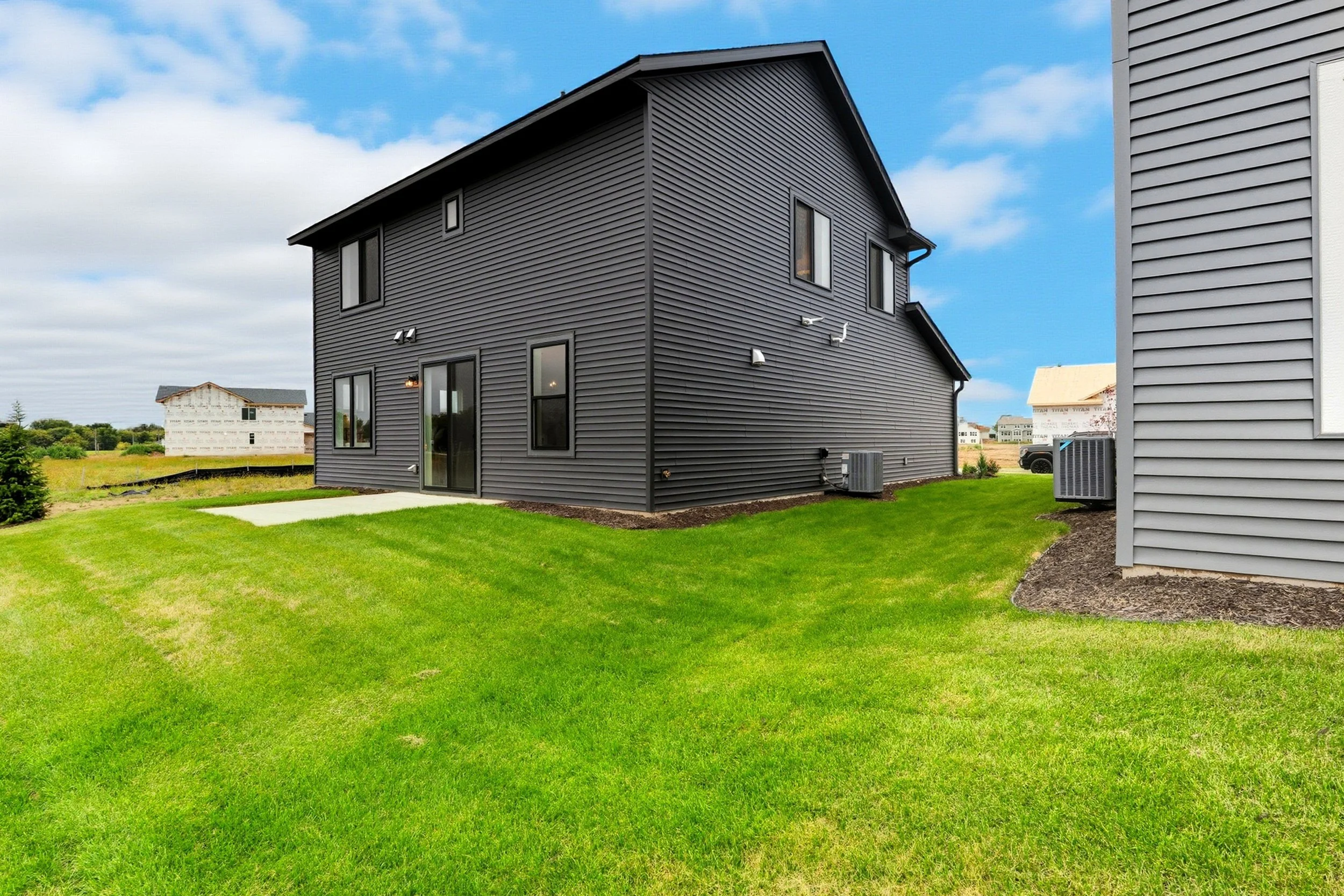 Backyard view of a black two-story house with green grass and a concrete patio, under a partly cloudy sky.