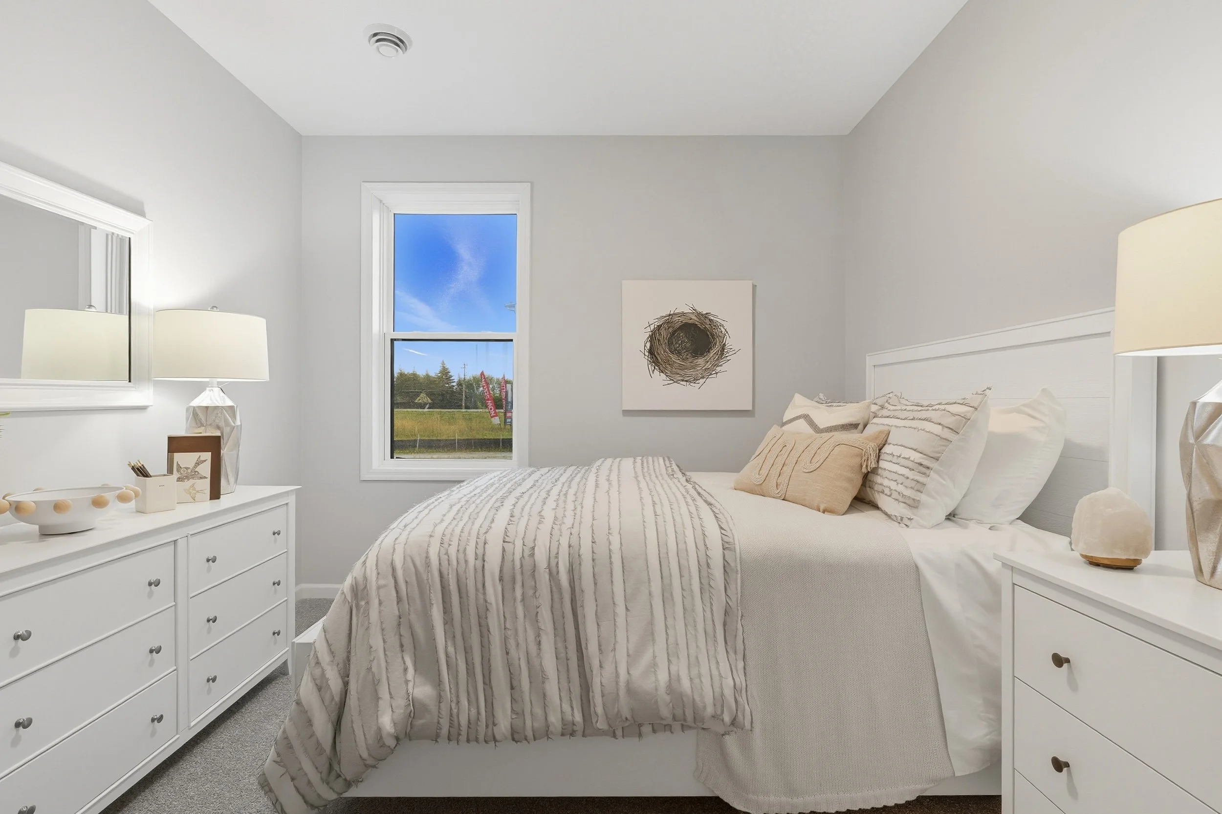 A bright bedroom with white walls and furniture, a bed with beige and white striped bedding, a window showing a blue sky and green landscape, and decorative items on nightstands.