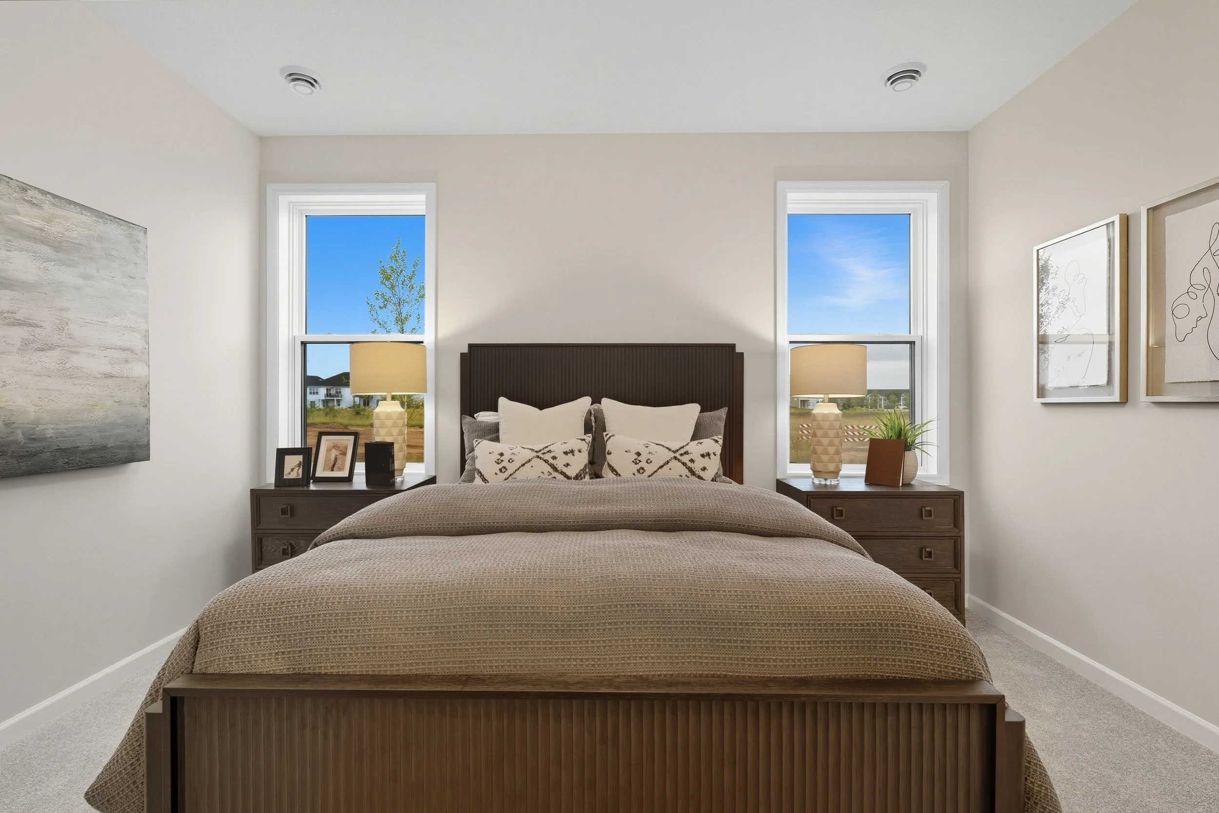 A neatly made bedroom with a large bed, two nightstands with lamps, framed photos, and decorative pillows, in front of two windows showing a view of a clear blue sky and green landscape.