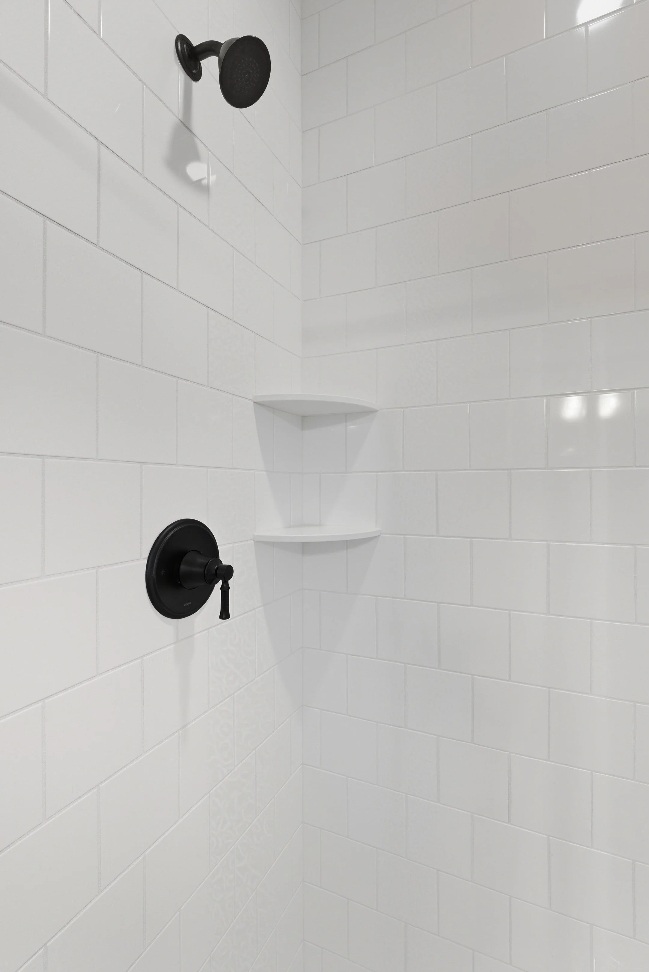 A clean, white tiled shower with a black showerhead and a black control handle, featuring built-in white corner shelves.