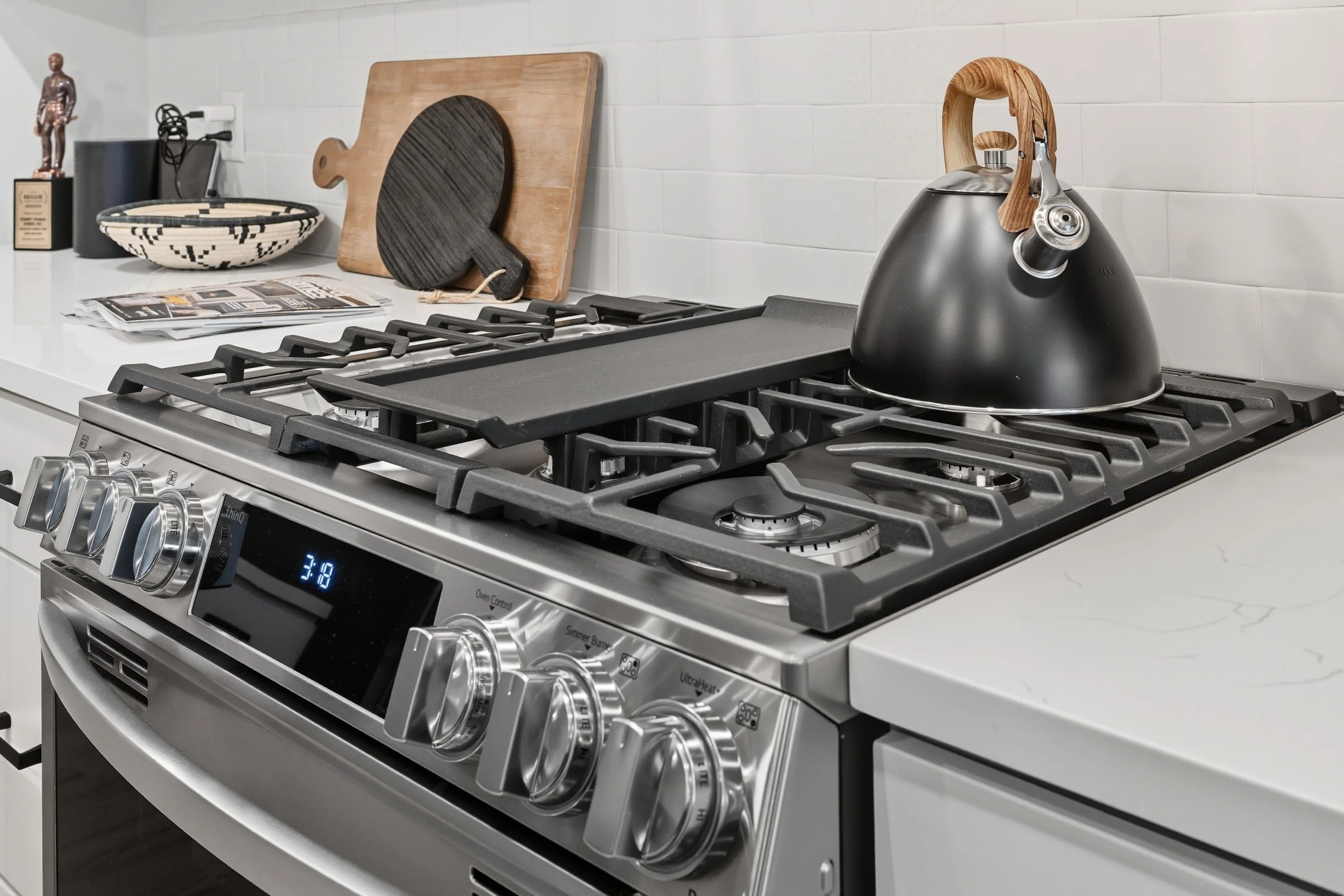 Modern stainless steel gas stove with a black kettle on top and kitchen countertop with cutting boards, a black-and-white bowl, and magazines.