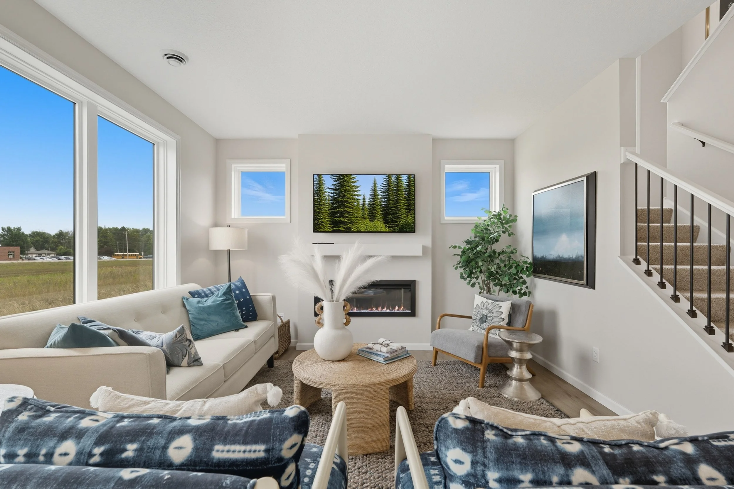 Living room with large windows, white walls, and modern furniture, including a white sofa with blue pillows, a gray armchair, a round woven coffee table with a decorative vase, wall-mounted TV, and a staircase.