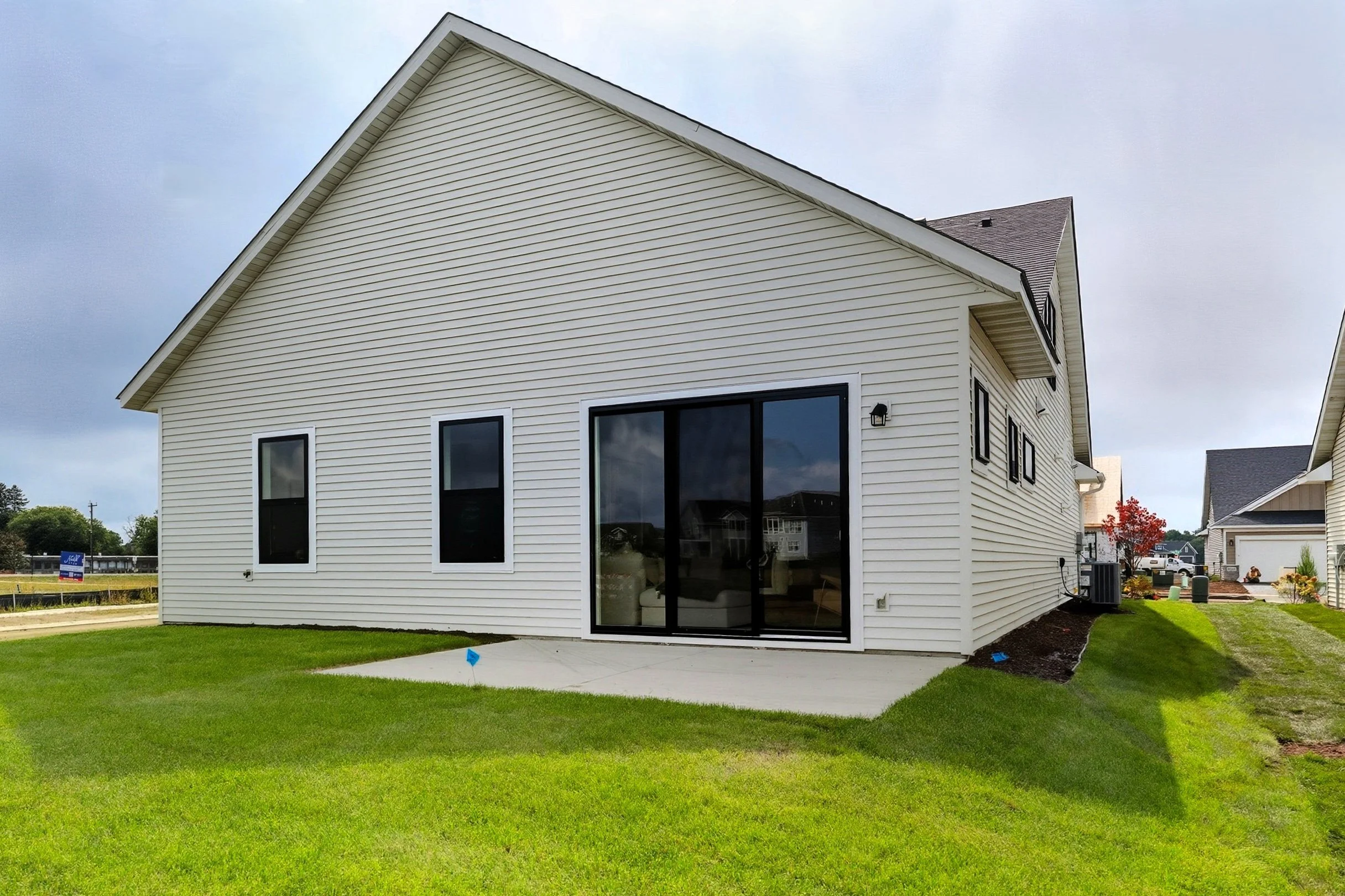 Rear view of a modern white house with black-framed windows and a sliding glass door, a concrete patio, green lawn, and neighboring houses in the background under a cloudy sky.