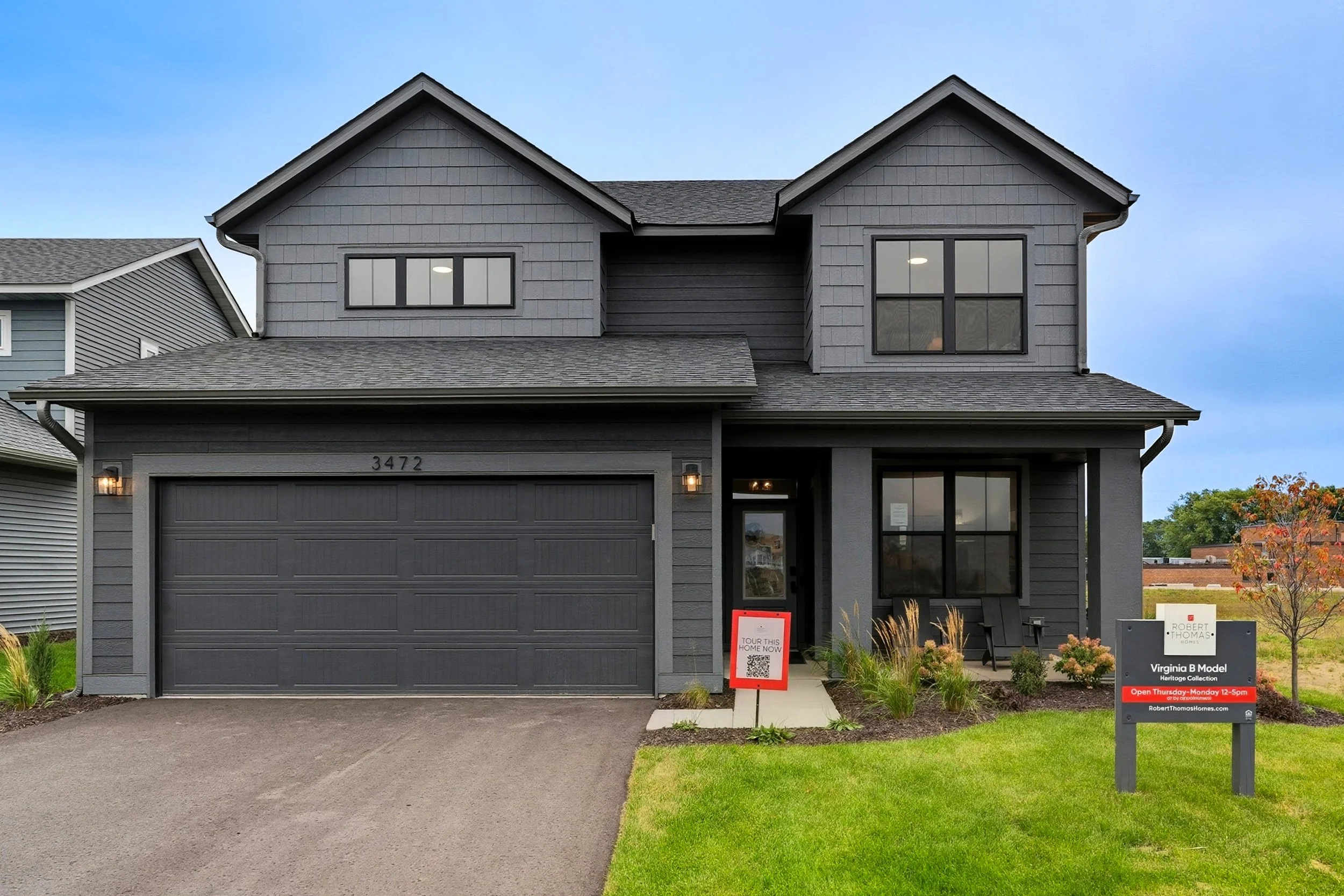 A two-story dark gray house with a garage, front porch, and a lawn. There are signs in front of the house indicating it's available for tours and open house hours.