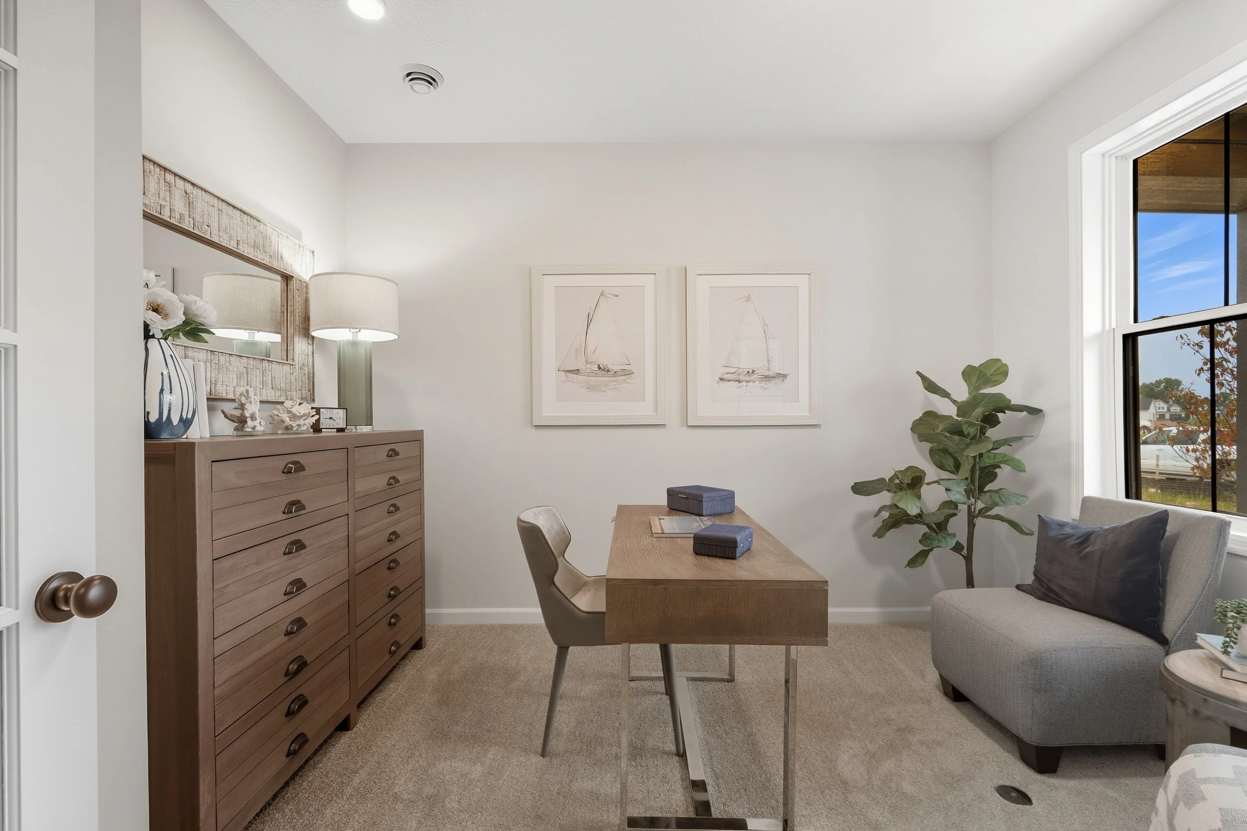 A modern, neutral-colored home office with a wooden desk, beige chair, gray armchair with a dark pillow, a tall dresser, a plant, and artwork of two sailboats on the wall.