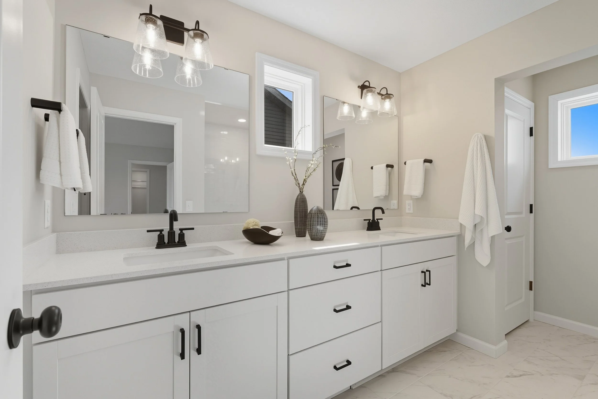 Modern bathroom with a long white vanity featuring two black sinks and faucets, large mirrors, black fixtures, white cabinets, and decorative vases with branches. There are two windows, one small and one larger, with white frames, and white towels ha
