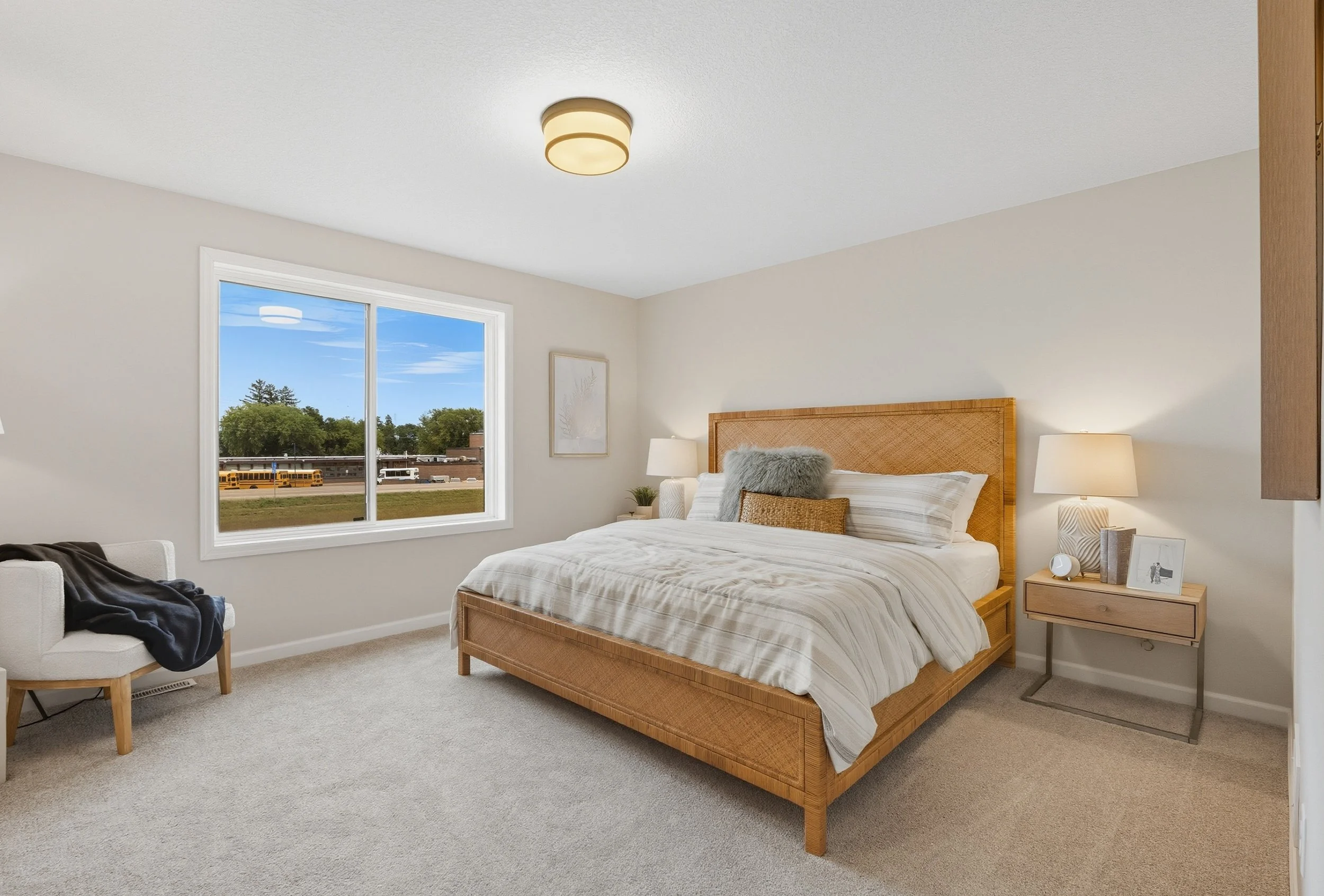 A bedroom with a large wooden bed, white and beige striped bedding, gray and brown pillows, two lamps on bedside tables, a framed artwork on the wall, a window showing an outdoor view, and a beige carpeted floor.