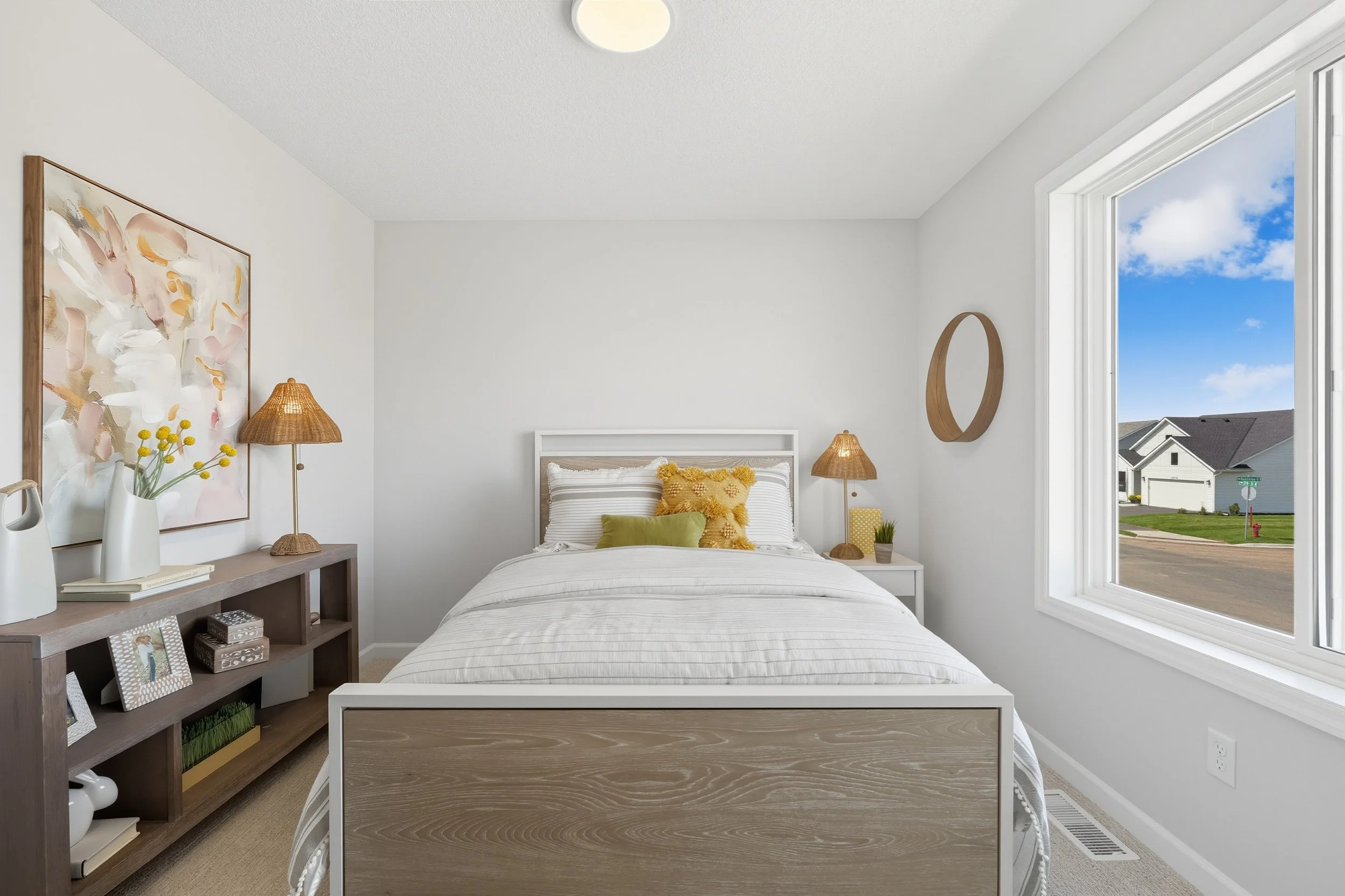 Well-lit bedroom with white walls, a bed with white and yellow pillows, a wooden frameless headboard, two bedside tables with lamps, a large window showing a street view, and a wooden shelf with photo frames, vases, and decorative items.