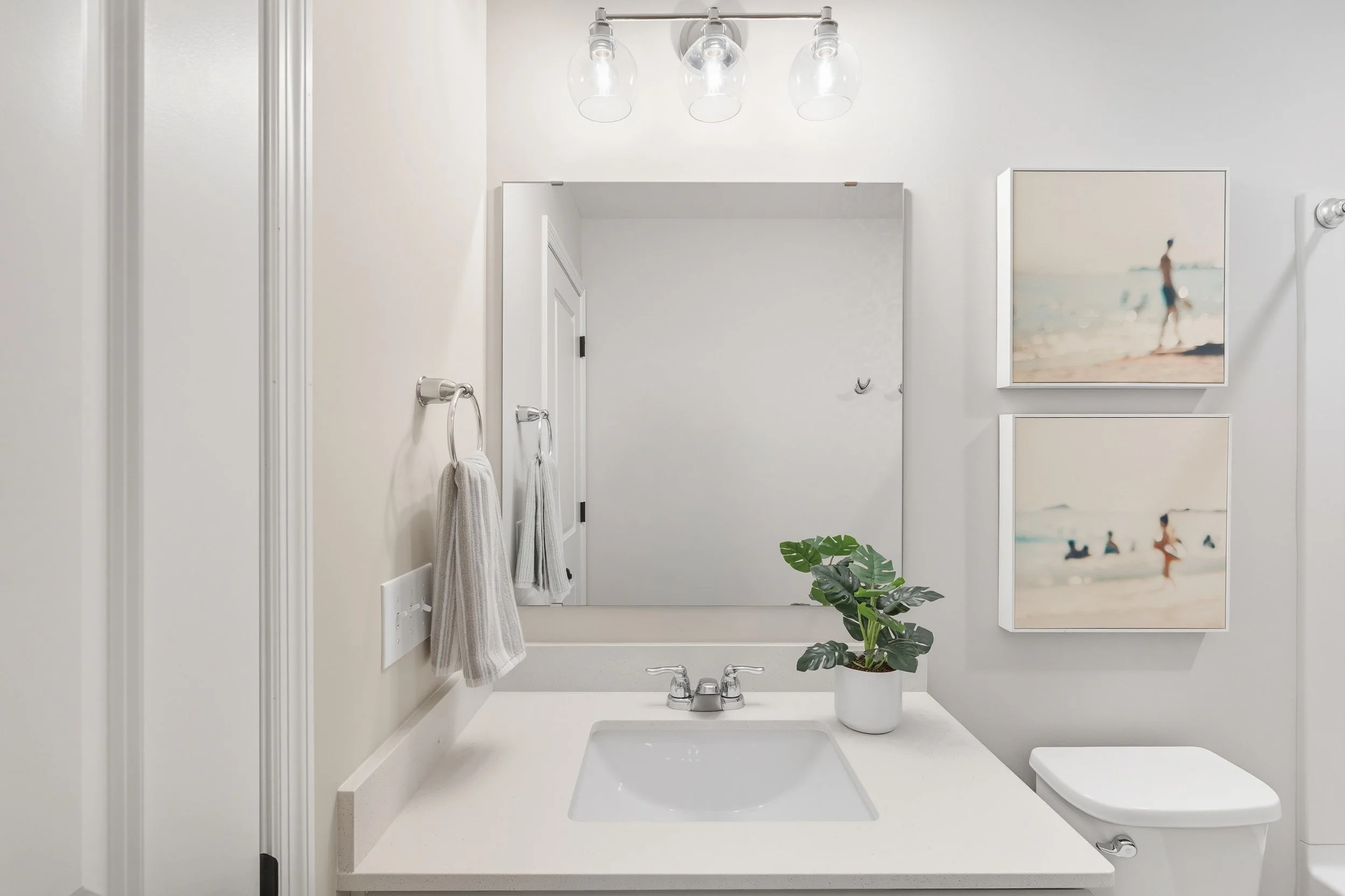 A clean, modern bathroom with a large mirror above a white countertop with a sink. There is a plant on the counter, two towels hanging on the wall, and two framed beach-themed photographs on the wall. The bathroom also has a toilet and three light bu