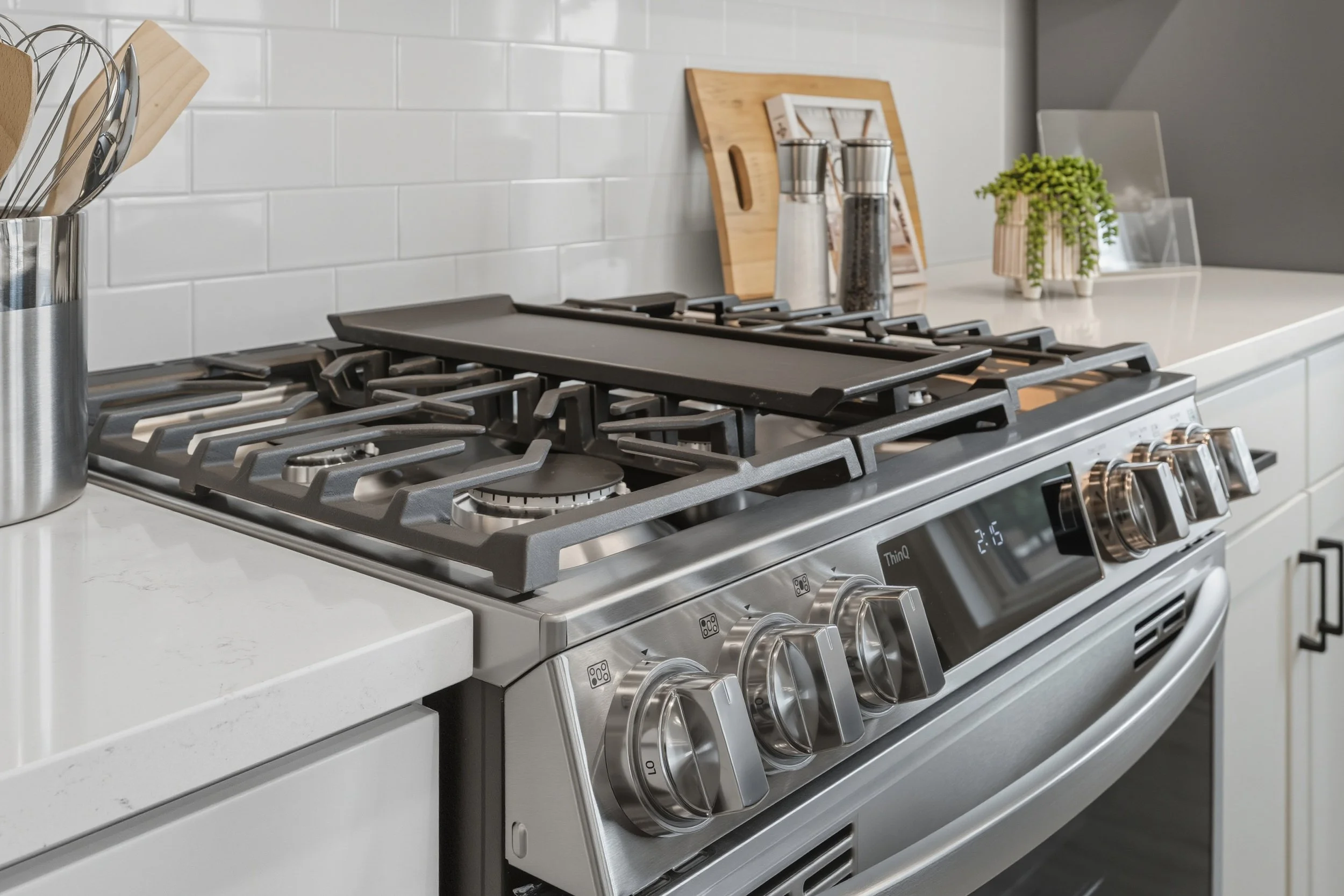 Modern stainless steel gas stove with black grates on a white kitchen countertop, with white subway tile backsplash and small decorative plants and kitchen accessories in the background.