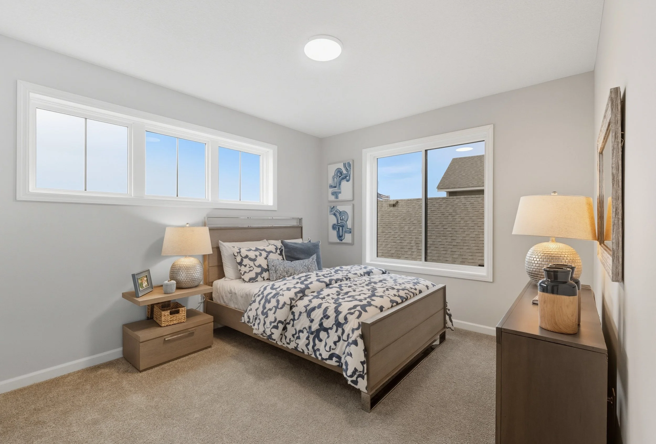 A bedroom with a bed, nightstand, and dresser. The bed has navy and white bedding, and two pillows. There are two lamps, one on each side of the bed. The room has two large windows with a view of rooftops and a clear sky.