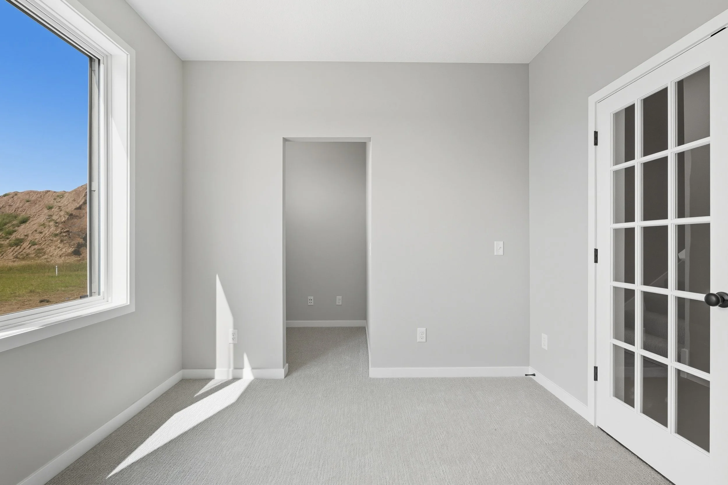 Empty room with gray walls, a window showing a daytime outdoor view, a glass door, and a small doorway leading to another room.