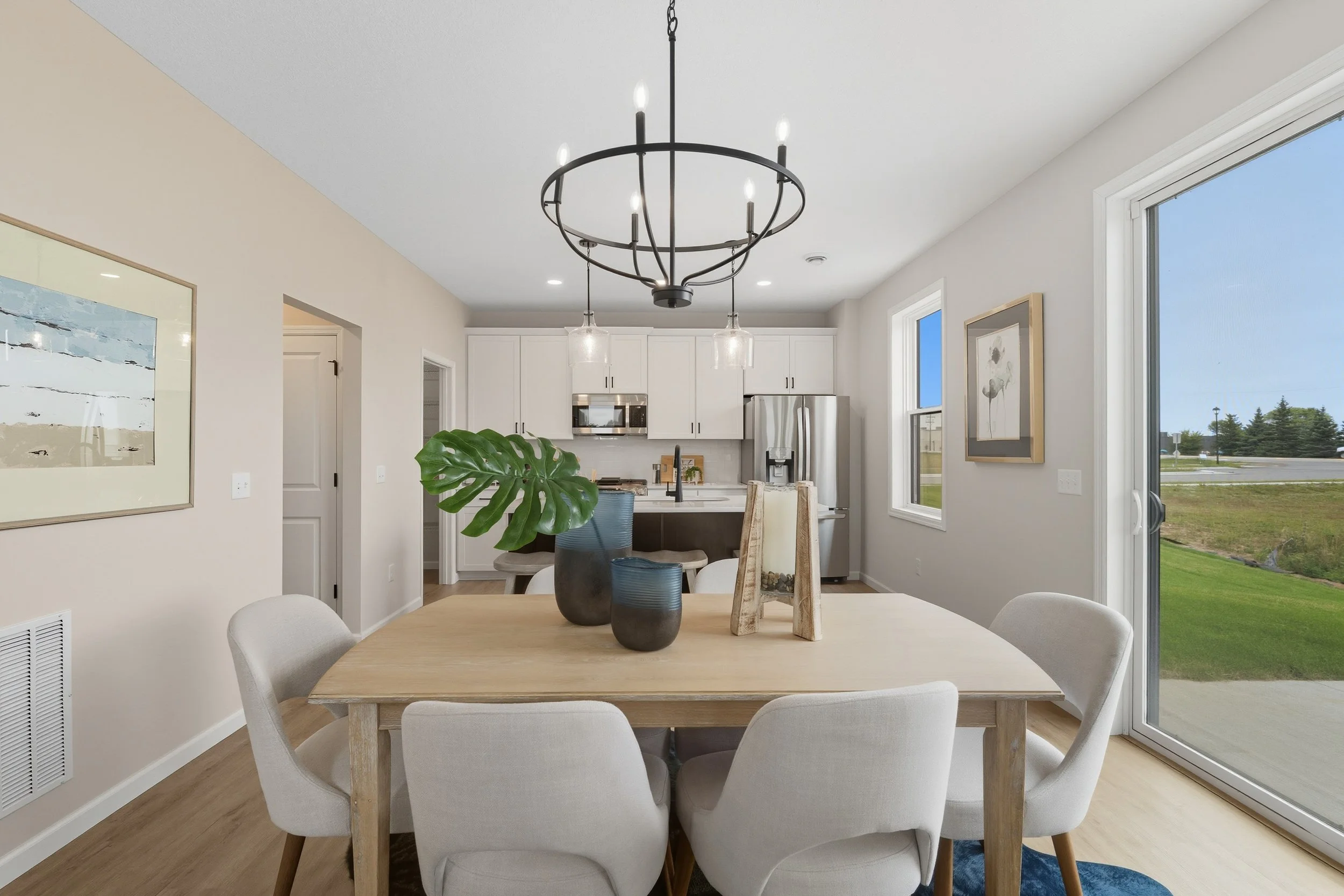 Dining area with round wooden table, six white chairs, large green leaf plant in black vase, modern chandelier, and a kitchen with white cabinets and stainless steel refrigerator in the background. Large sliding glass door opens to outdoor view.