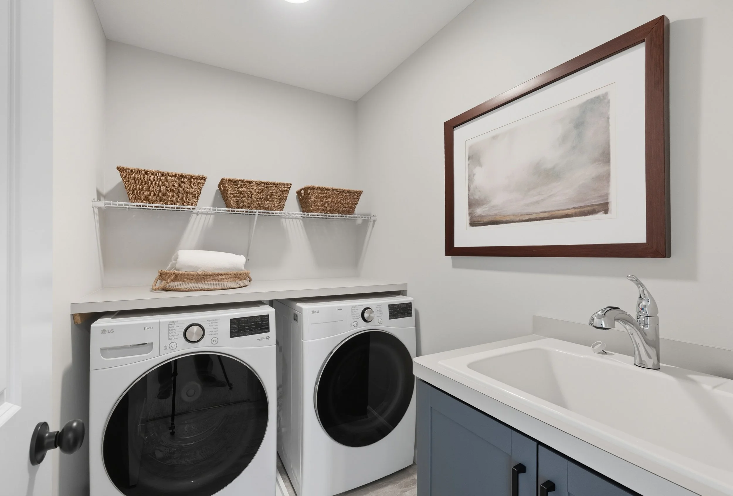 A laundry room with a front-loading washing machine and dryer, a white utility sink, a framed landscape painting, and baskets on a wire shelf.