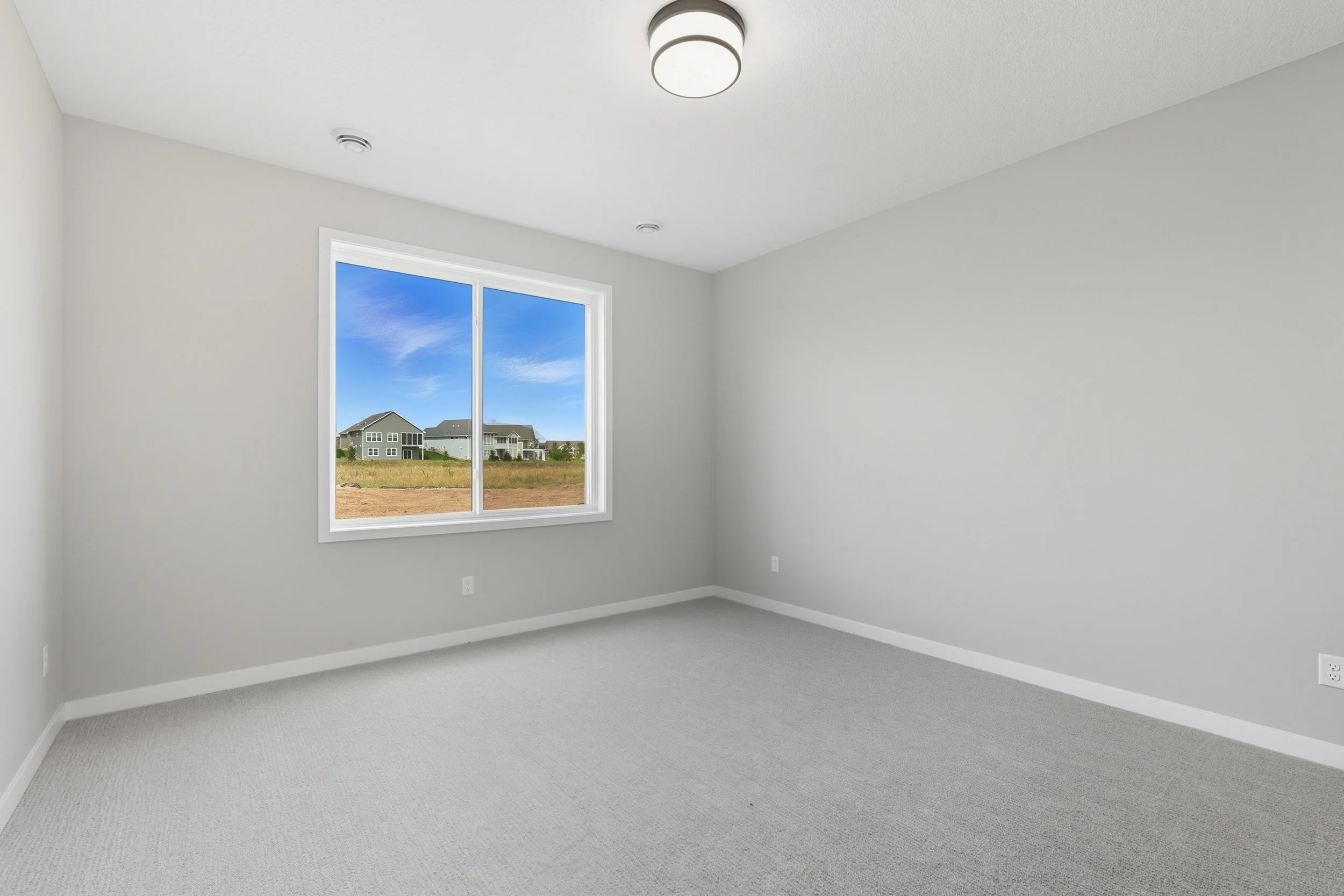 Empty room with light gray walls, gray carpet, modern ceiling light, and a window showing houses and a blue sky outside.