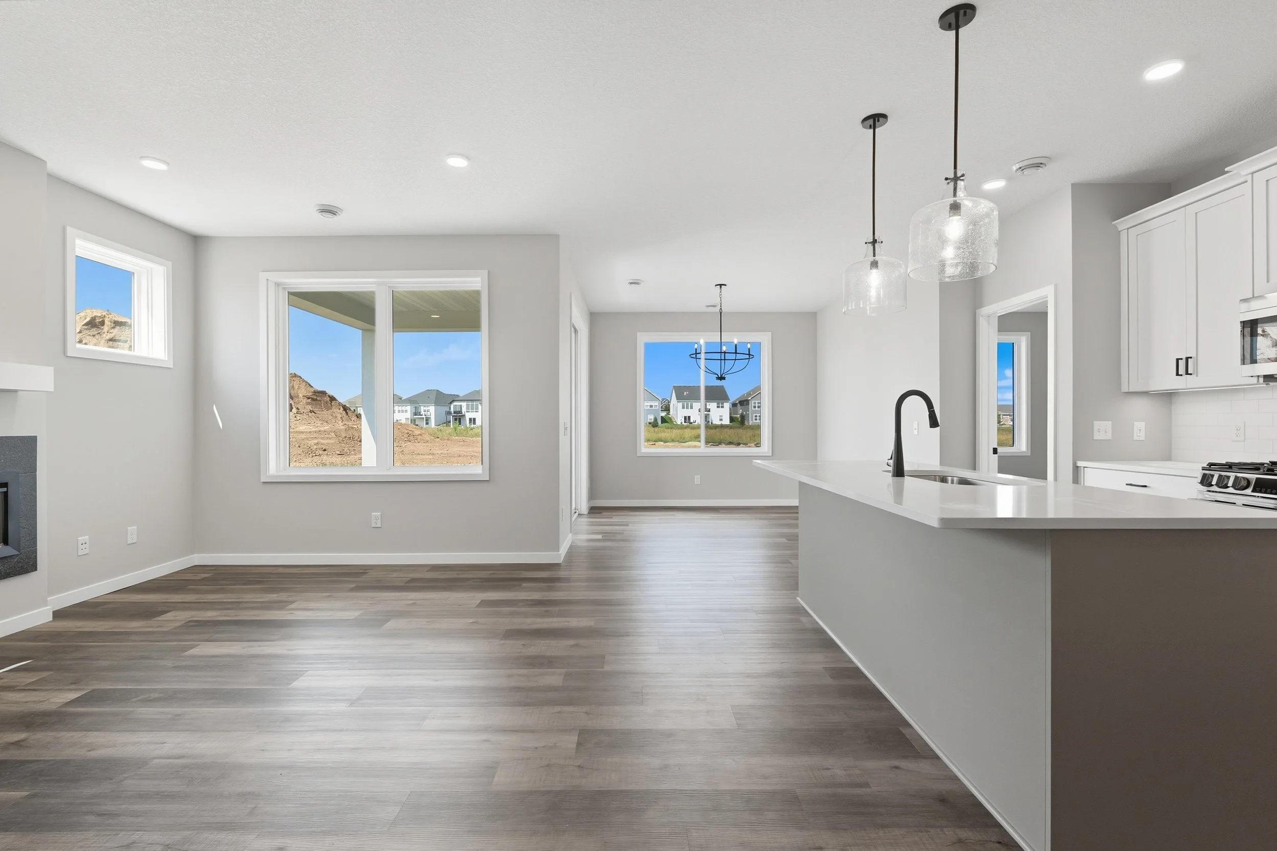Empty modern kitchen and dining area with large windows, light gray walls, white cabinetry, and island counter, with view of houses and mountains outside.