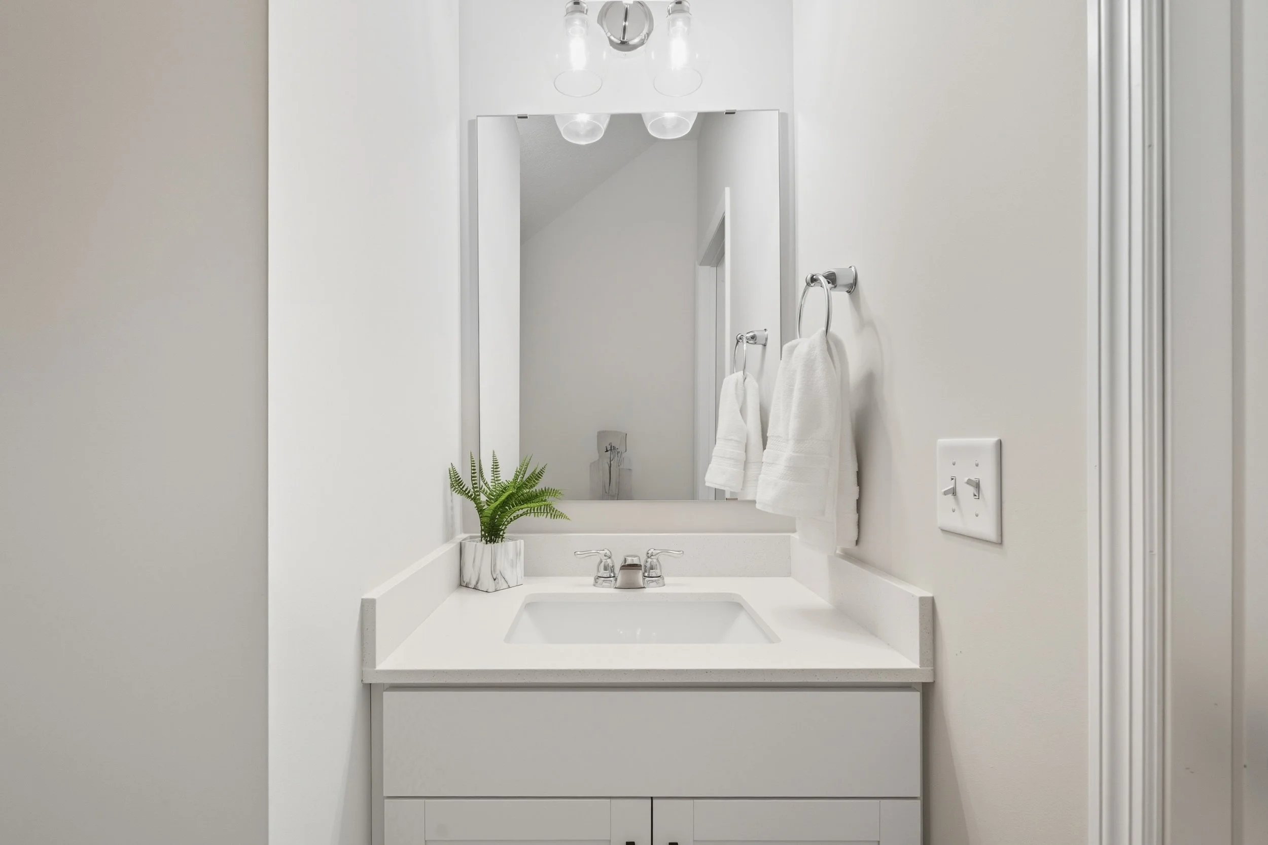 A clean, white bathroom vanity with a rectangular sink, a mirror, a small potted plant, and two white towels hanging on the wall, with lighting fixtures above the mirror.