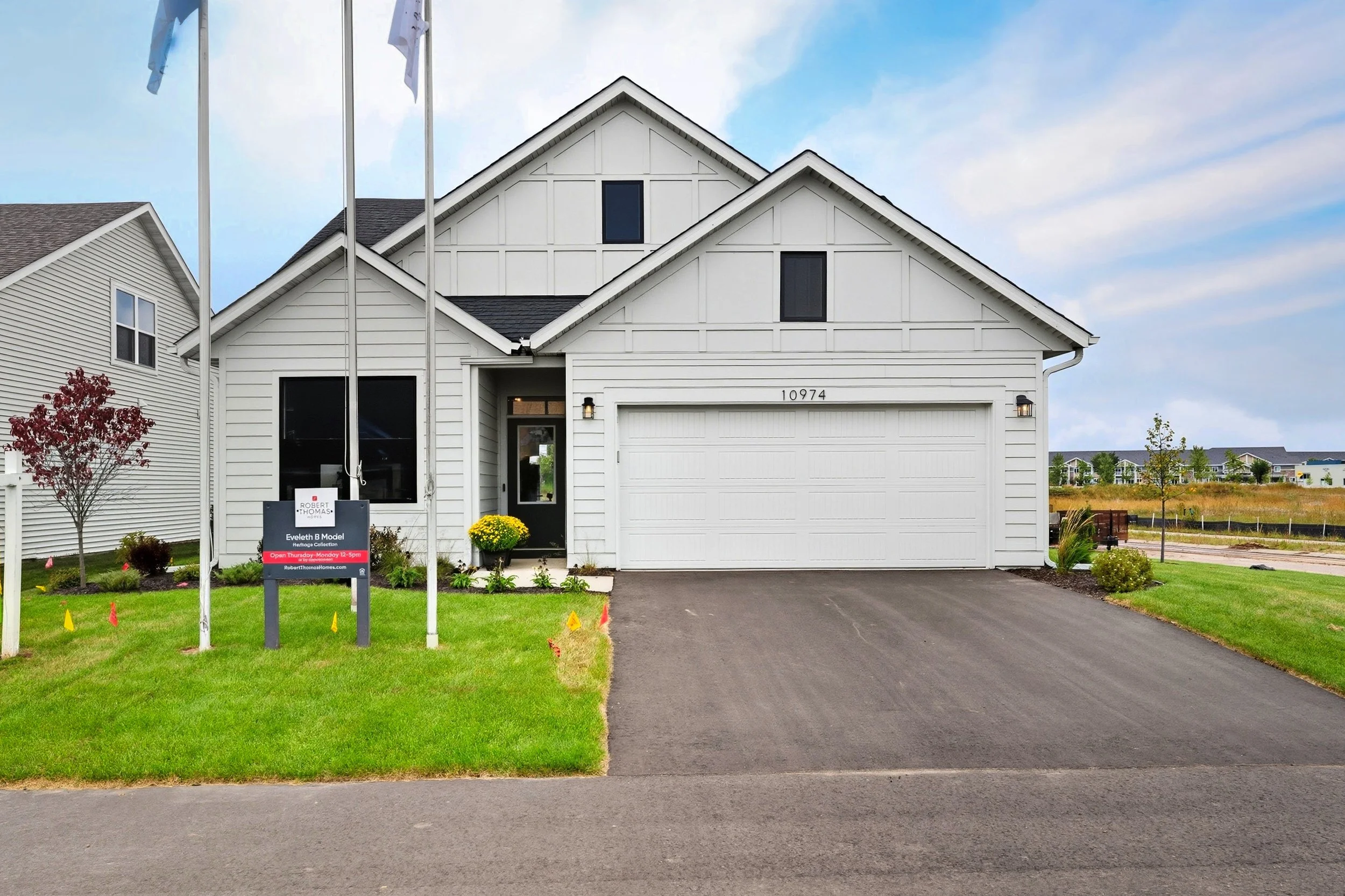A white two-story house with black roof, in a new neighborhood, with a green lawn, a black driveway leading to a garage, and a sign in front with real estate information. There are flagpoles in the yard, and a small tree with red leaves on the left.