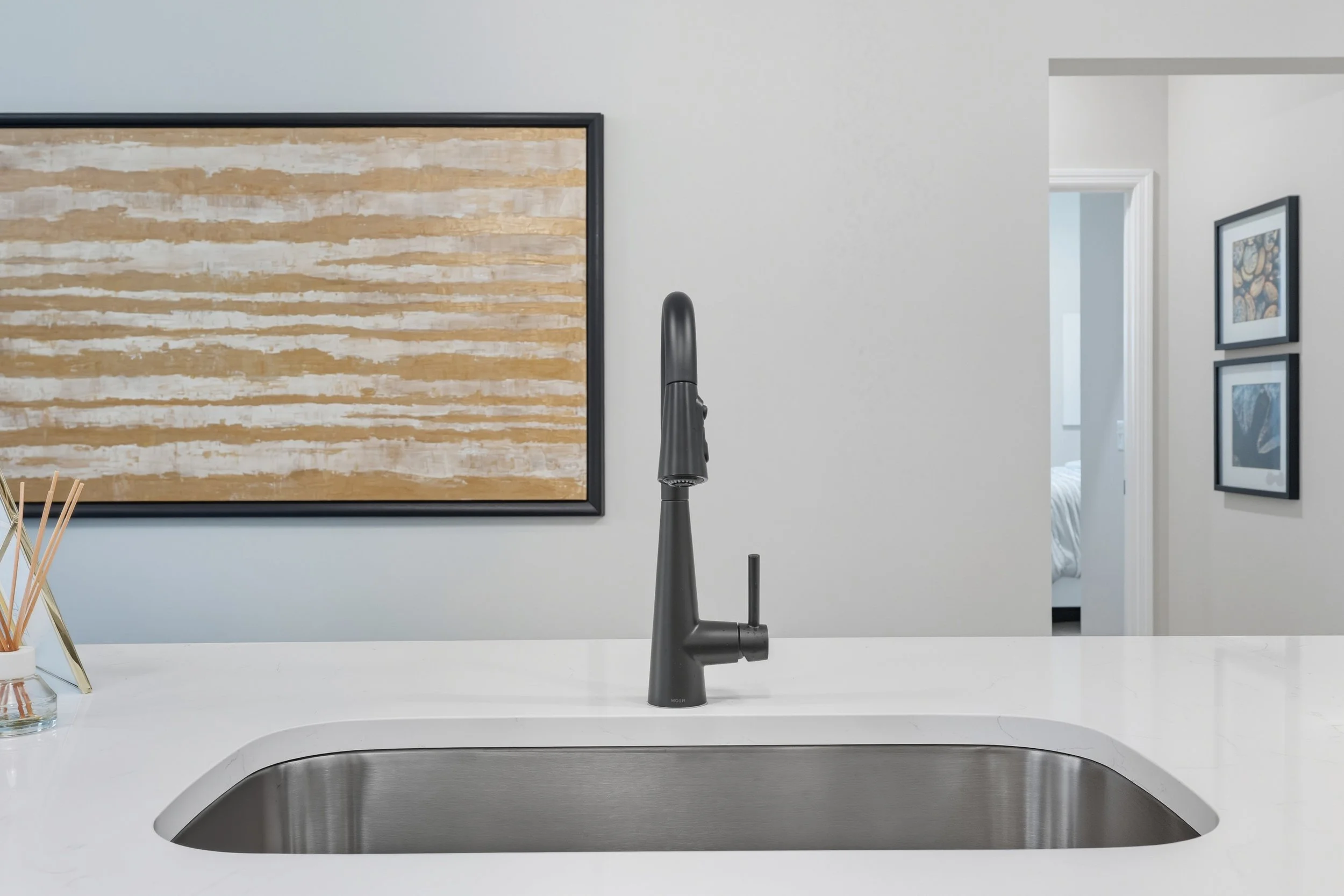 Modern kitchen sink with black faucet on white countertop, with abstract wall art and framed photos in the background.