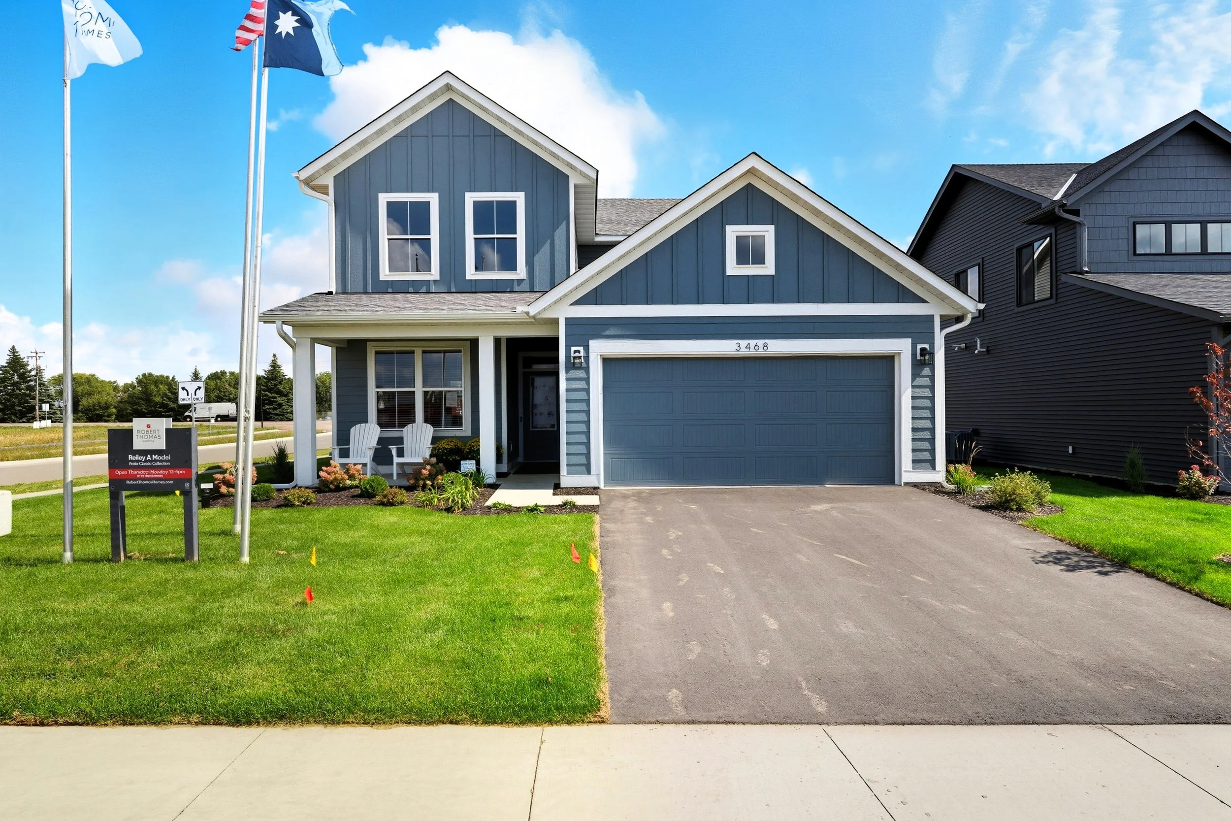 A two-story blue house with white trim, a front porch with two white chairs, a green lawn, a driveway, and three flagpoles in front. The house has the number 3468 on it.