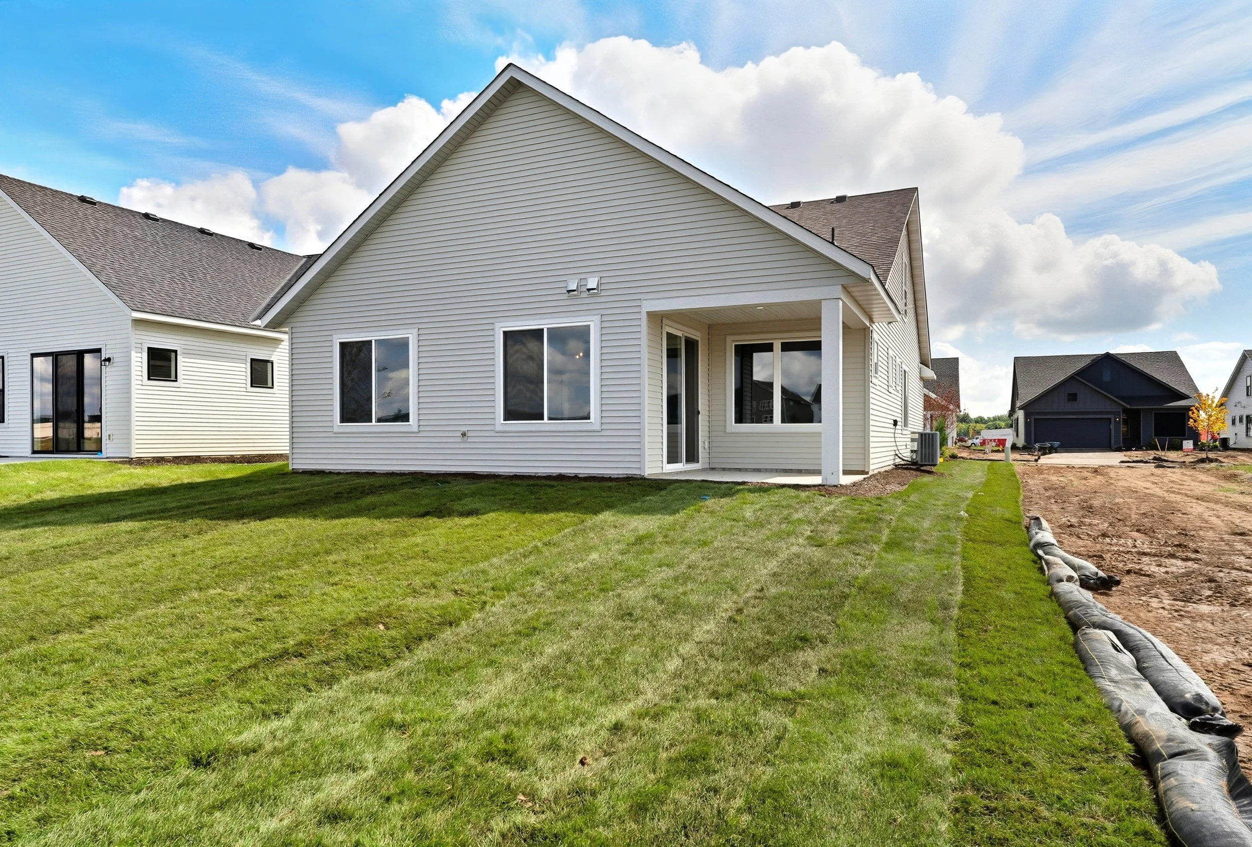 Newly built white house with a backyard lawn, a small covered patio, and neighboring houses under a partly cloudy sky.