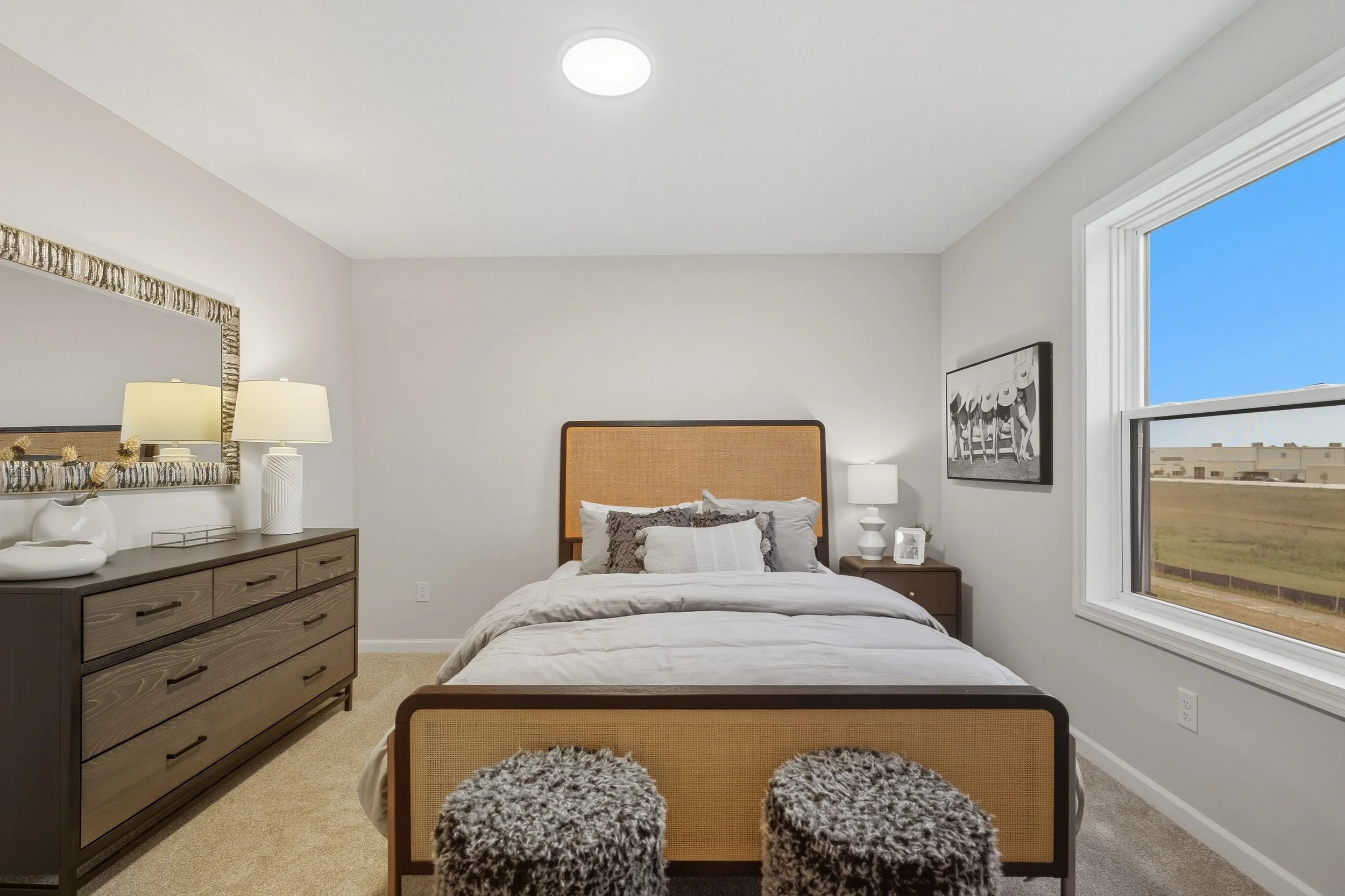 Bedroom with a wooden bed frame and beige headboard, white bedding, dark gray and white pillows, dark brown nightstand with a lamp, framed black and white artwork on the wall, large window showing outside view of a grassy field and buildings, beige c