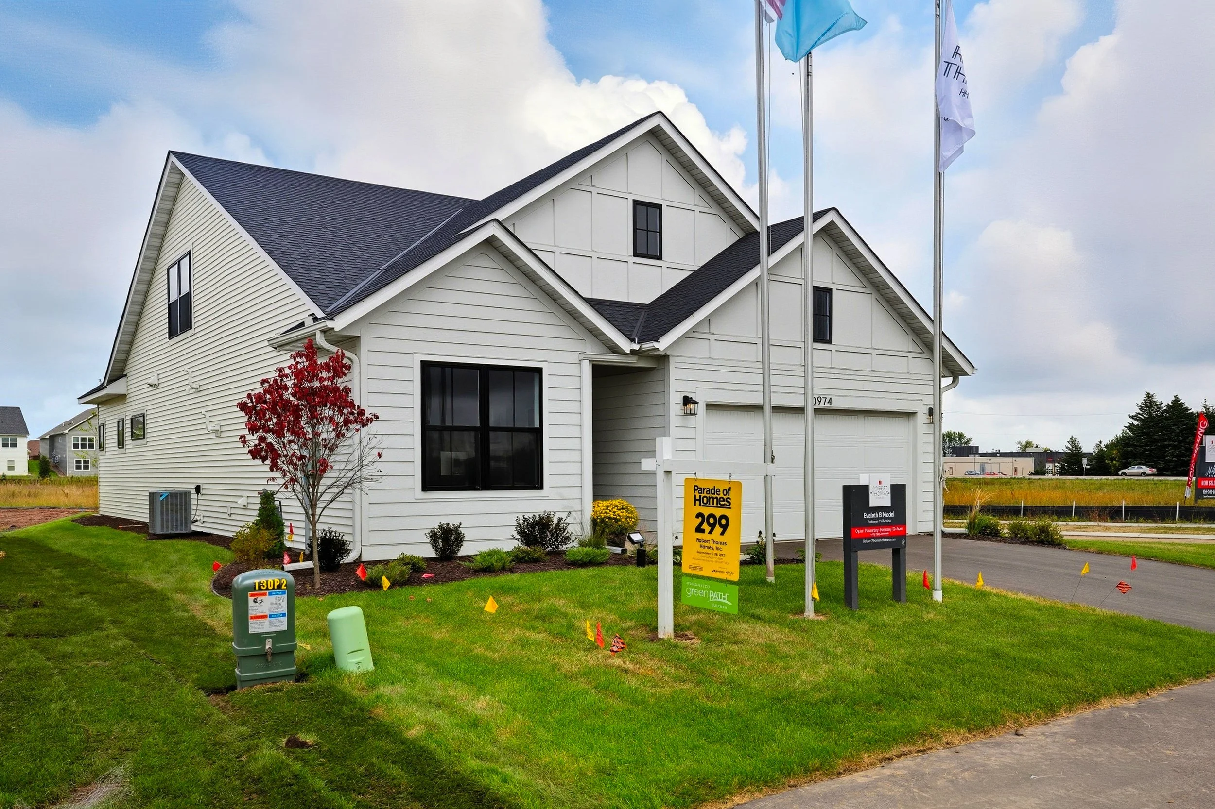 New white house with black windows and a double garage, landscaped yard with a small red tree, flags, and a sign indicating price of $299,000 for homes for sale.