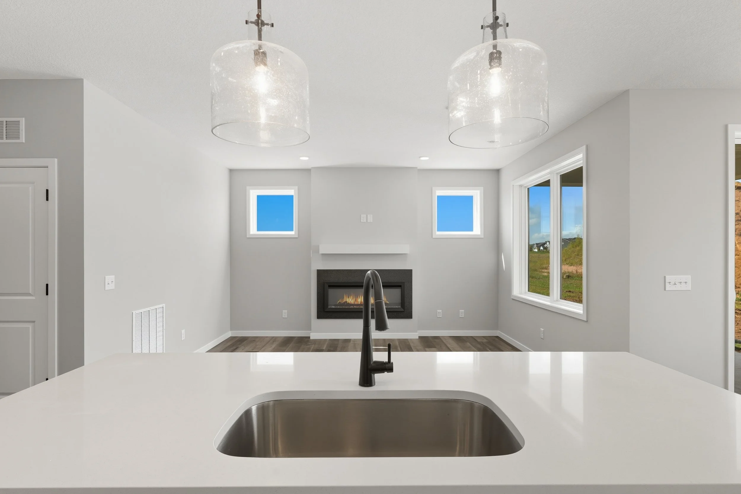Modern kitchen with white island countertop and a black faucet, overlooking a living room with a fireplace, two small high windows, and large sliding glass doors showing a green outdoor landscape.