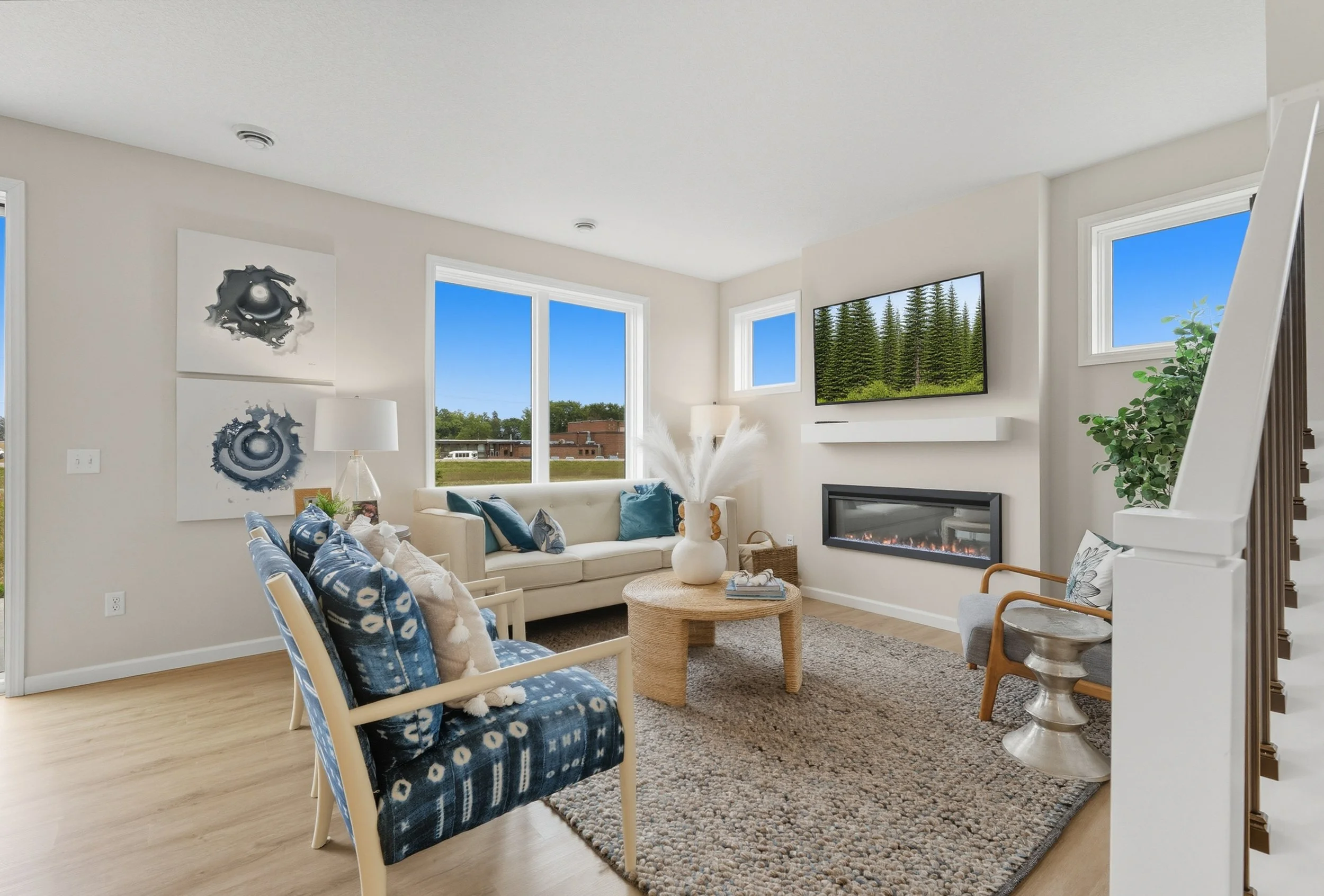 Living room with white walls, large windows, a wall-mounted TV, a fireplace, blue and white cushions, a coffee table with decorative vase, indoor plant, artwork, and light wood flooring.